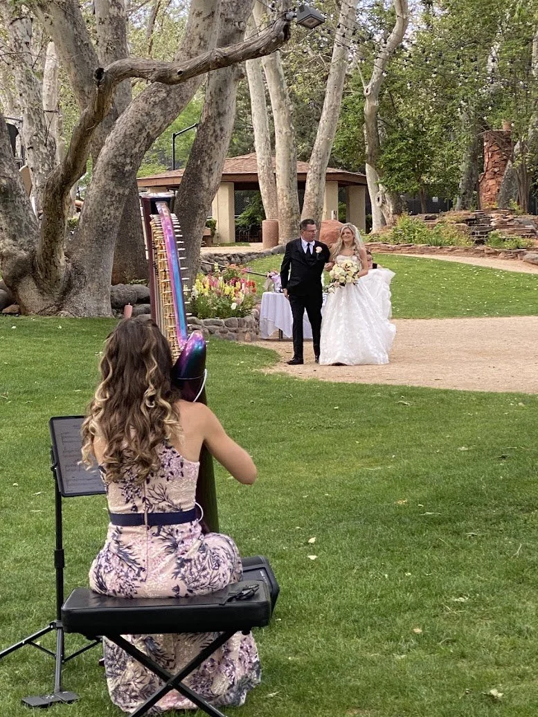 A woman playing a large harp outdoors near a river with trees in the background. Harpist for Sedona, Arizona Weddings. Sedona Elopement. Live music for Elopement. 