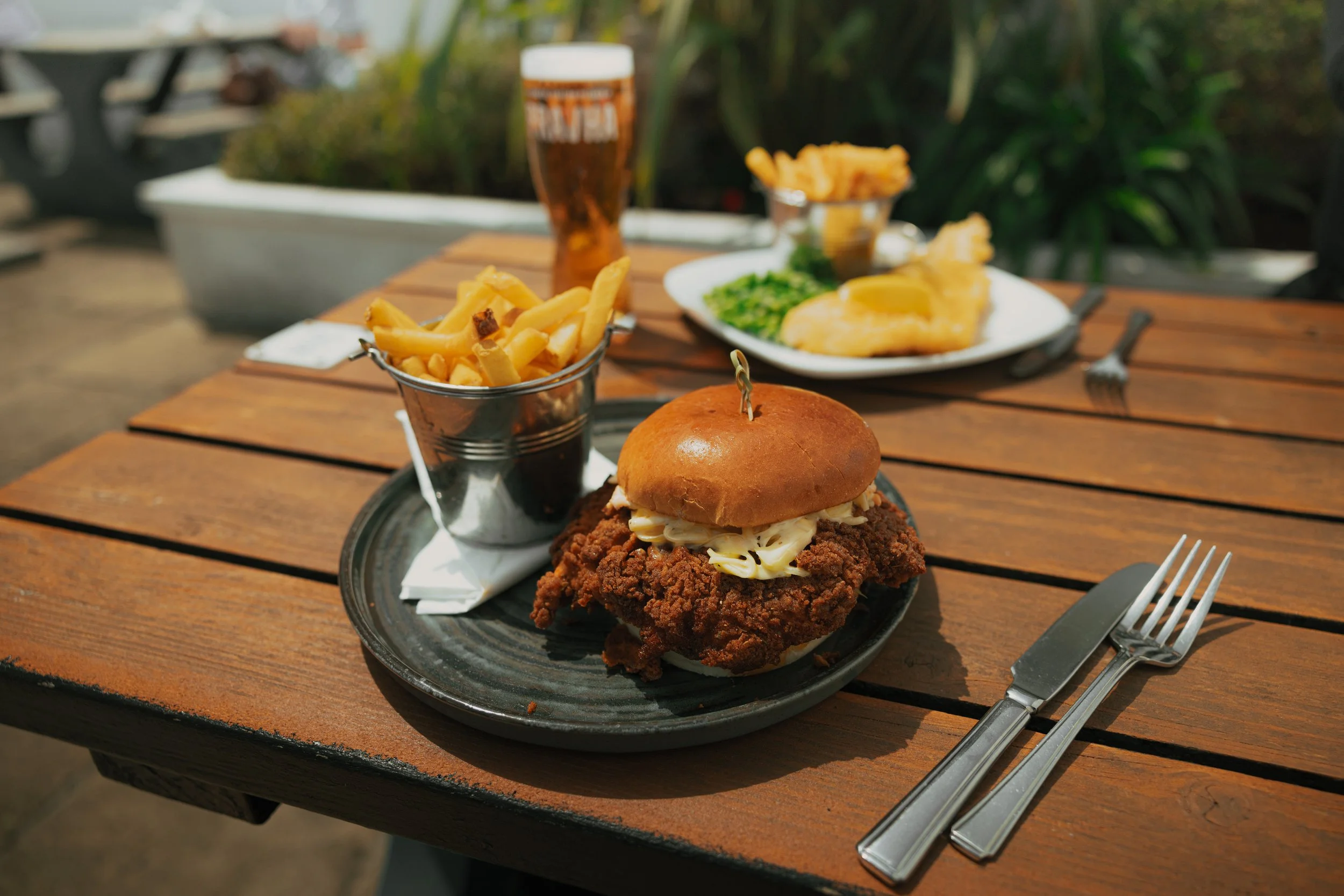 A burger and chips, with a pint of beer and fish & chips in the background on an outdoor bench