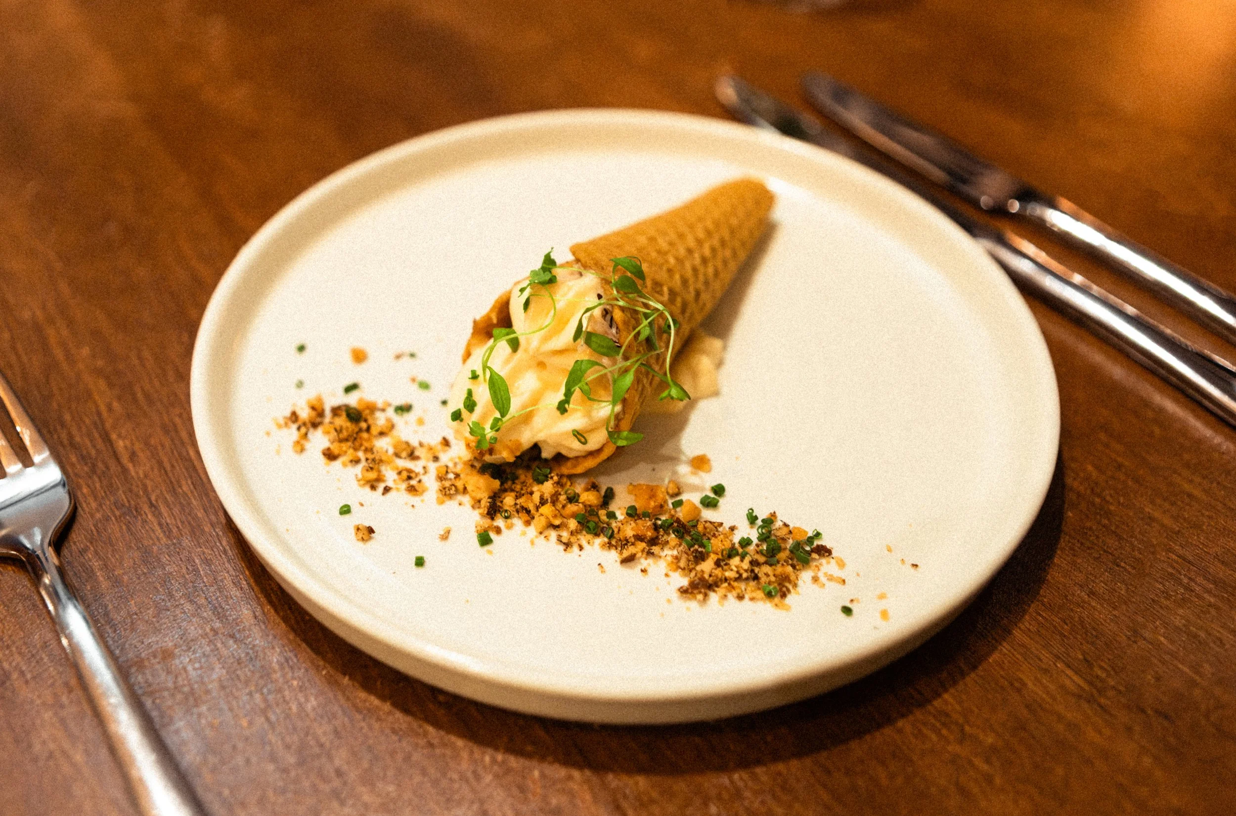 A plated dish featuring a piece of meat topped with greens and purple edible flowers, served with a side of chickpeas in tomato sauce, on a rustic ceramic plate.