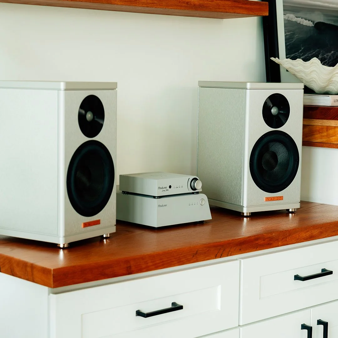 Two white Magico bookshelf speakers with black driver cones are placed on a wooden surface, alongside a silver Wattson Audio  amplifier & Streamer