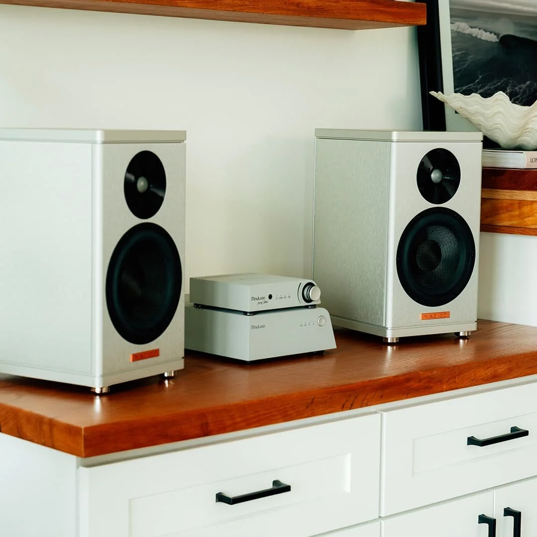 Two white Magico bookshelf speakers with black driver cones are placed on a wooden surface, alongside a silver Wattson Audio  amplifier & Streamer