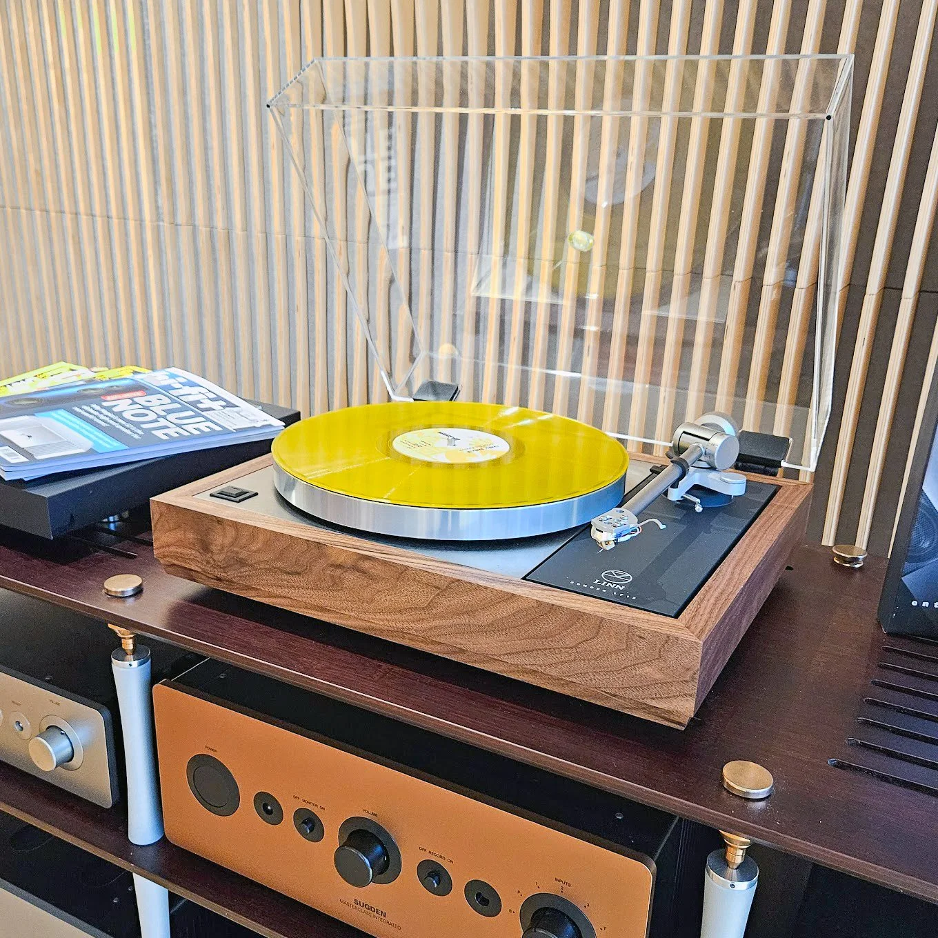 A Linn LP12 Klimax turntable with Bedrok Plinth & Radikal Power Supply, with a yellow vinyl record on it, with a transparent dust cover, positioned on a dark wooden hifi rack.