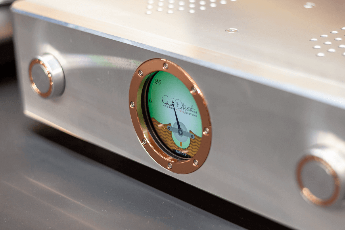 Close-up of a silver Pendulum with a green analog input level meter in the center, featuring a signature and the text 'Master Audio Systems.' Two round knobs are visible on either side of the meter.