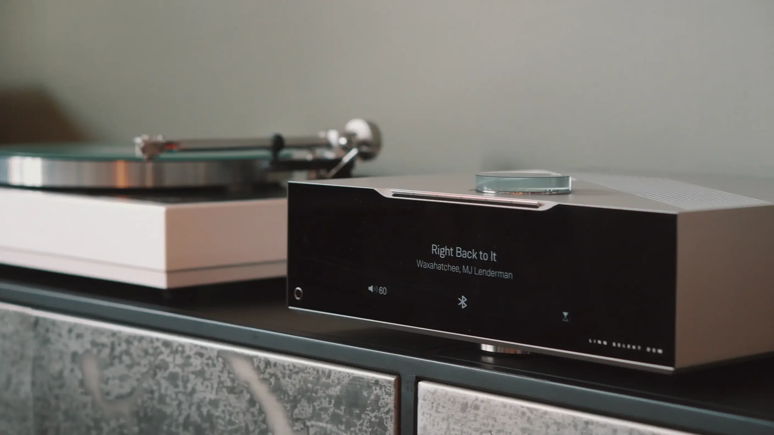 A Linn Selekt Music System in a minimalistic setting. In the foreground, a black device with a digital screen displaying song information and controls is visible.