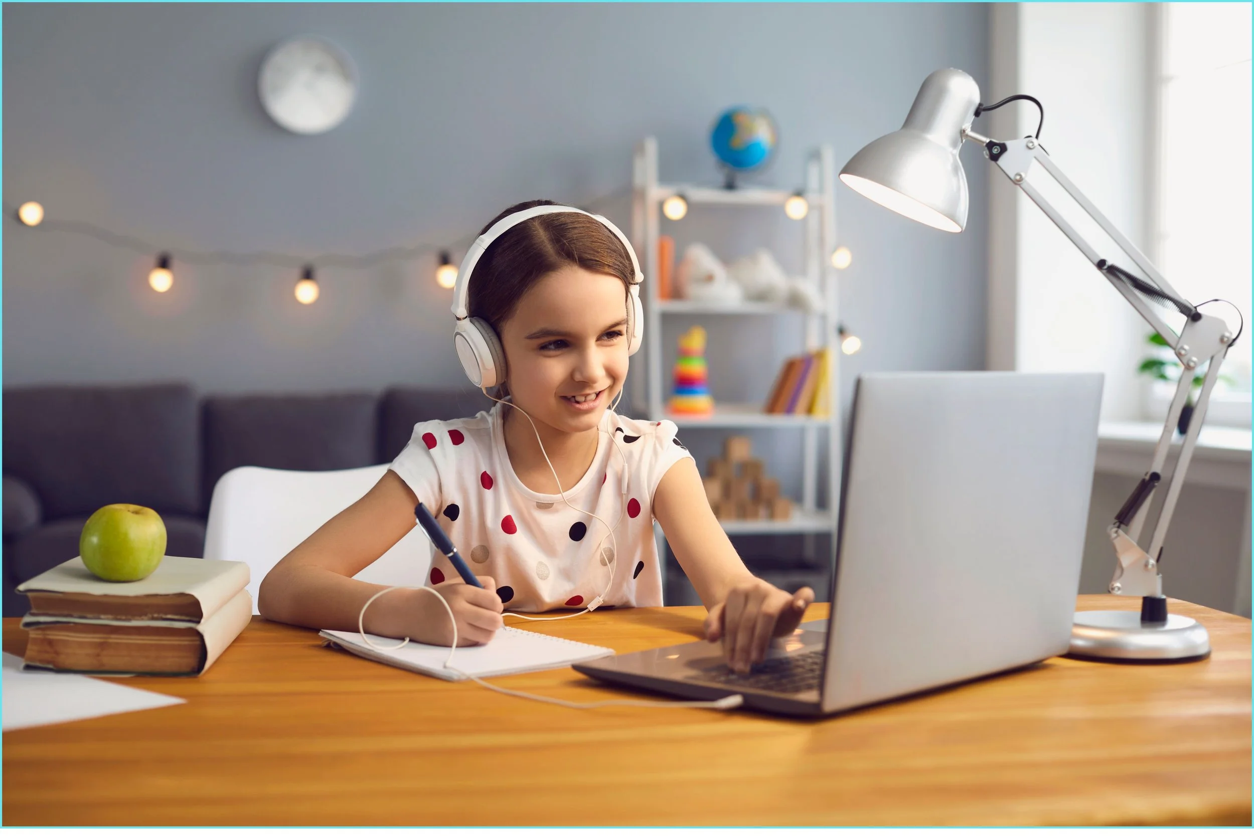 A young girl sitting at a desk with a laptop, wearing headphones, writing in a notebook, with a green apple and books on her left, and a modern desk lamp on her right, in a cozy room with a bookshelf and string lights in the background.