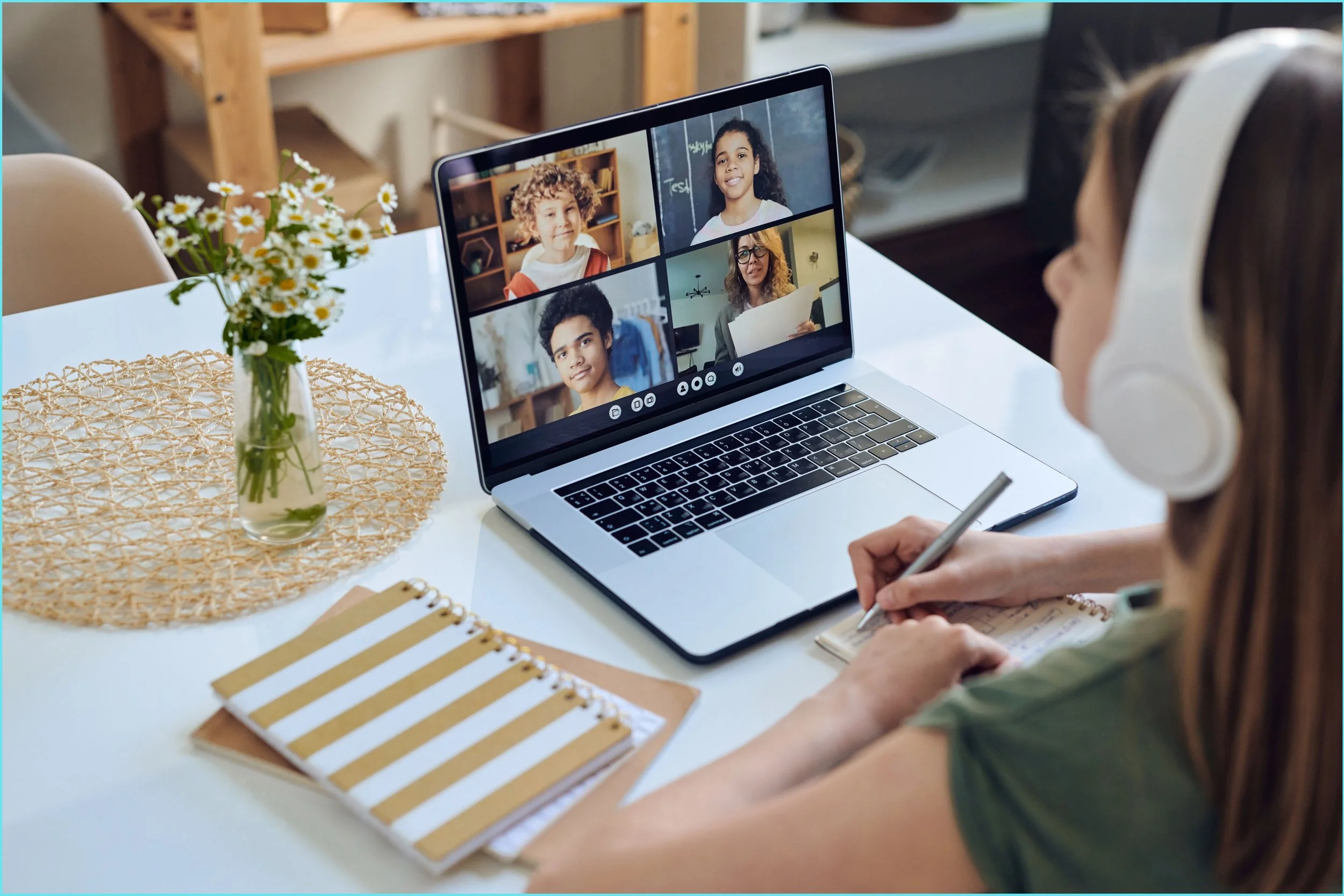 A woman wearing headphones and taking notes is participating in a virtual meeting with five children on her laptop screen, in a room with a vase of daisies, a striped notebook, and a woven placemat on the table.