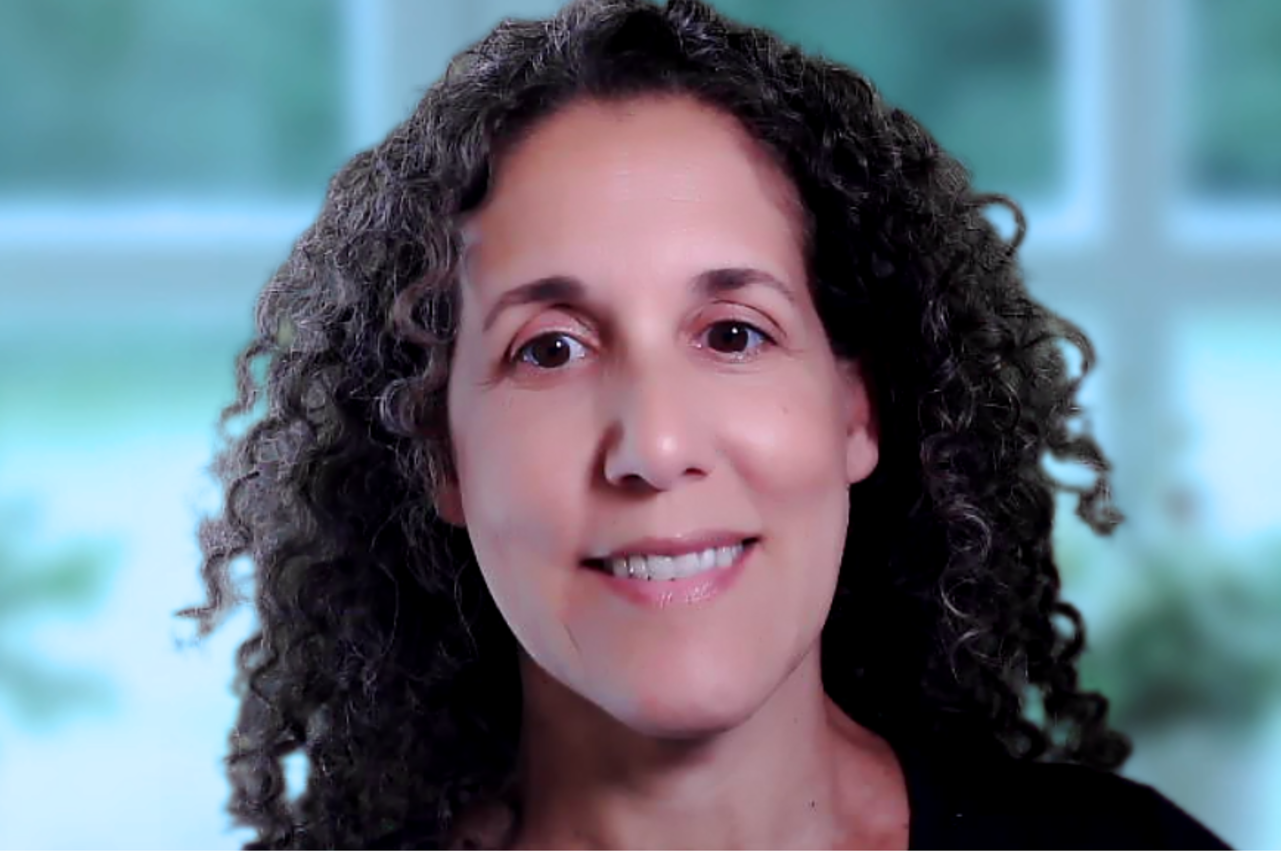 A woman with curly dark hair and fair skin smiling softly, with a blurred window and outdoor scenery in the background.
