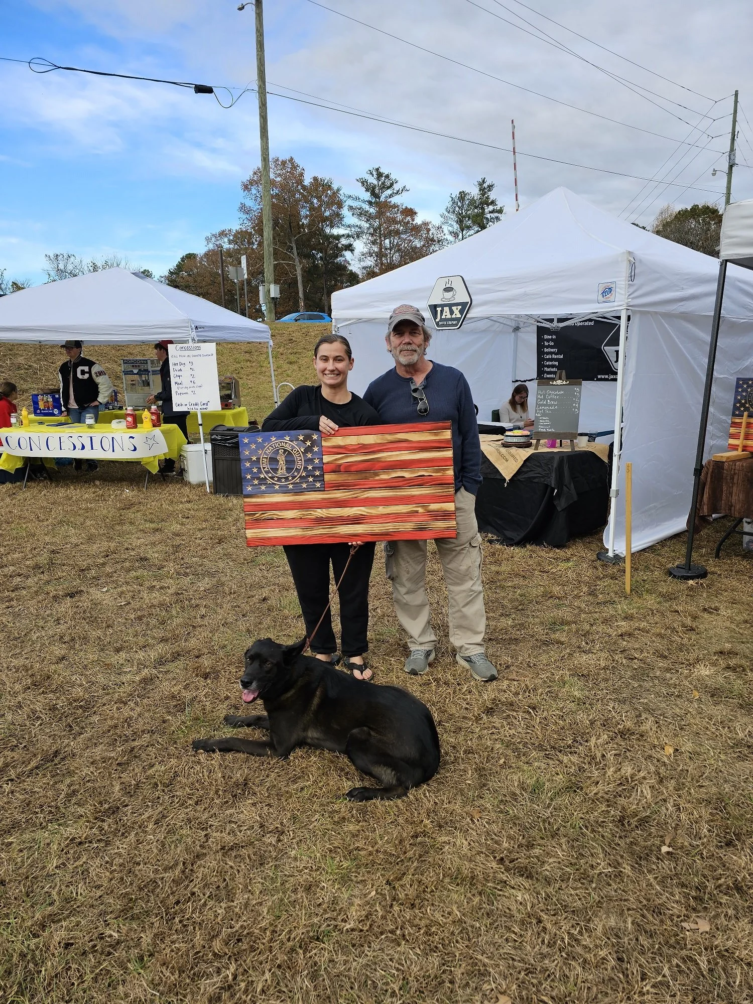 Two people standing outdoors under tents, holding a wooden American flag design, with a black dog lying on the grass in the foreground.