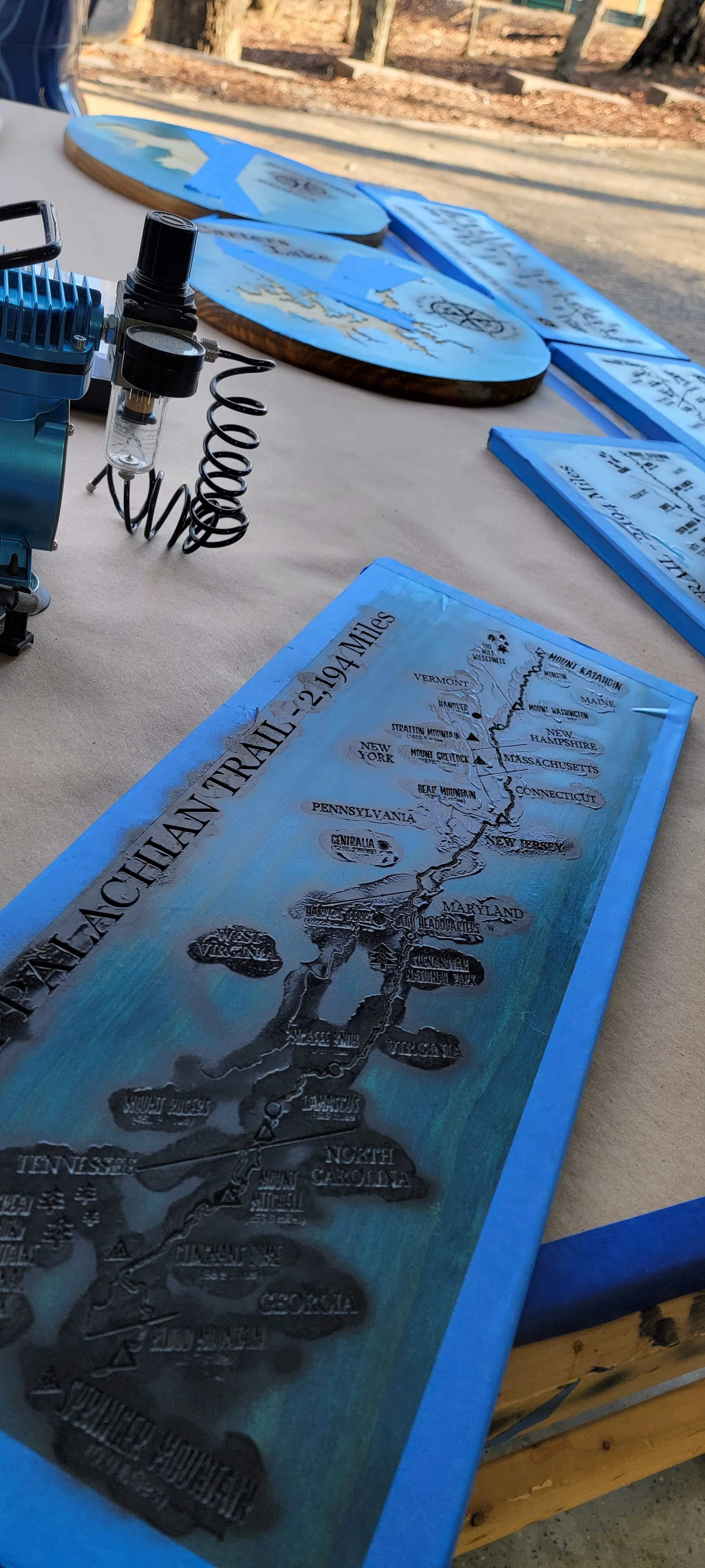 Wooden plaques depicting the Appalachian Trail map with a paint sprayer nearby, on a workbench.