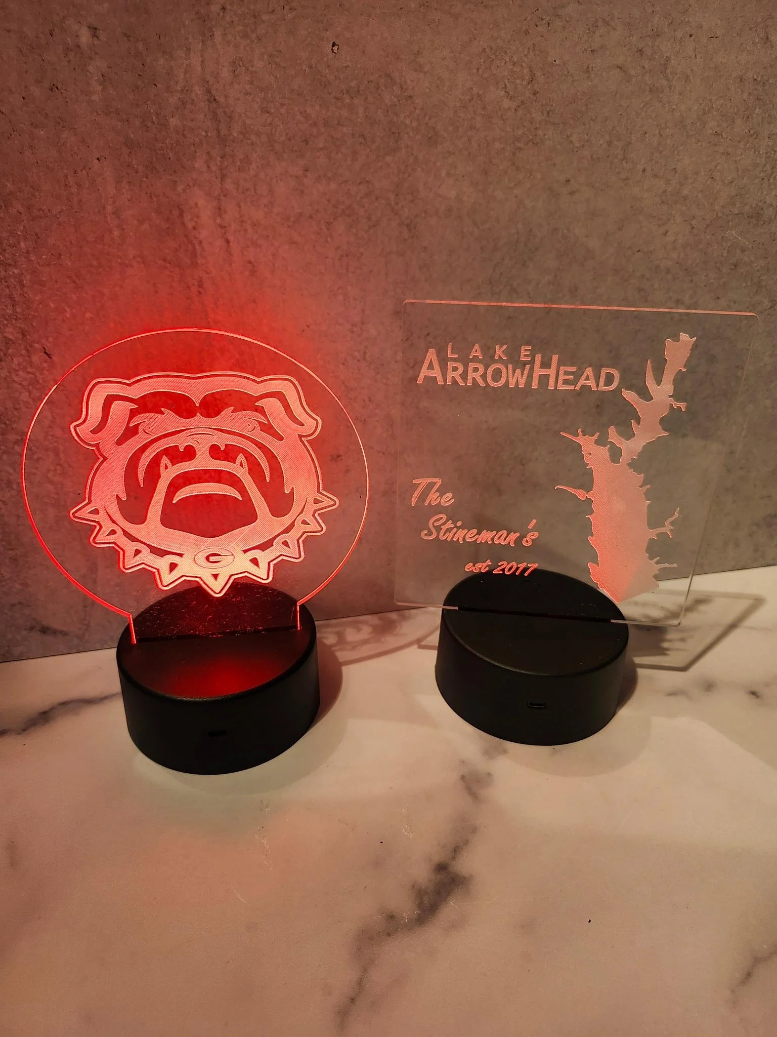 Two decorative LED acrylic plaques with stands on a marble surface. The left plaque features a stylized bulldog's face illuminated in red. The right plaque displays the words 'Lake Arrowhead' with a map outline and the text 'The Stineman's est 2017.'
