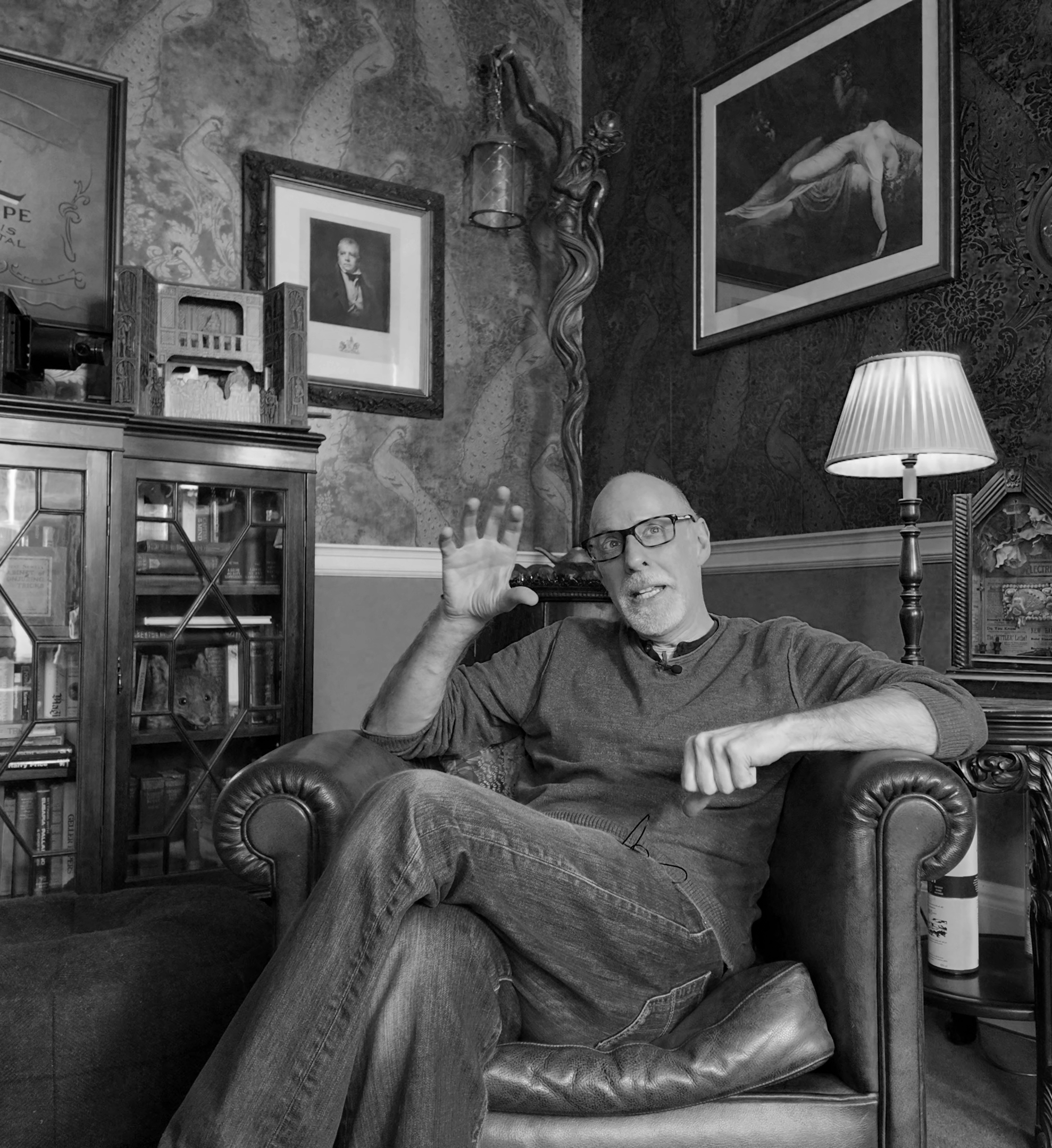 A man with glasses and a beard, sitting on an armchair in a room decorated with framed artwork and patterned wallpaper, waves with his right hand, with a lamp and a dark cabinet in the background.