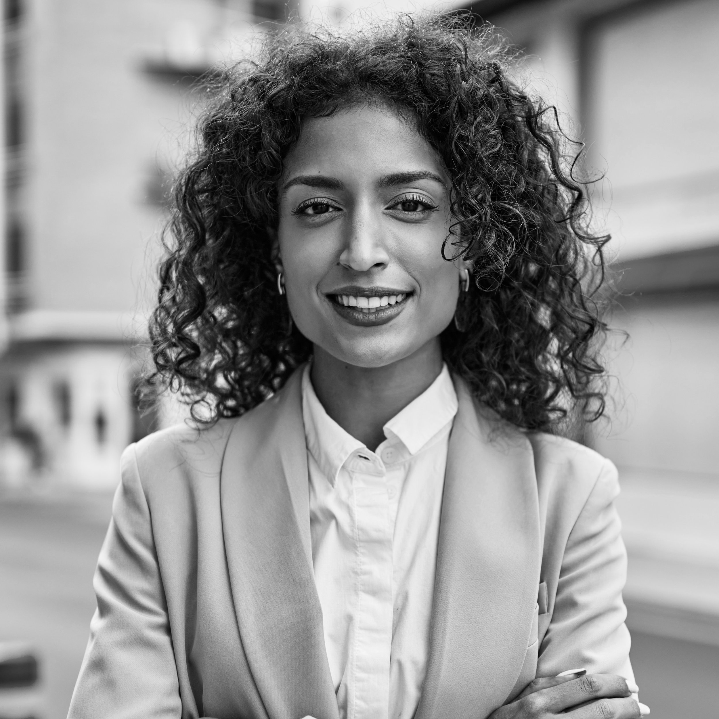 Black and white portrait of a smiling woman with curly hair, wearing a light-colored blazer and a white shirt, standing outdoors in front of a blurred city background.