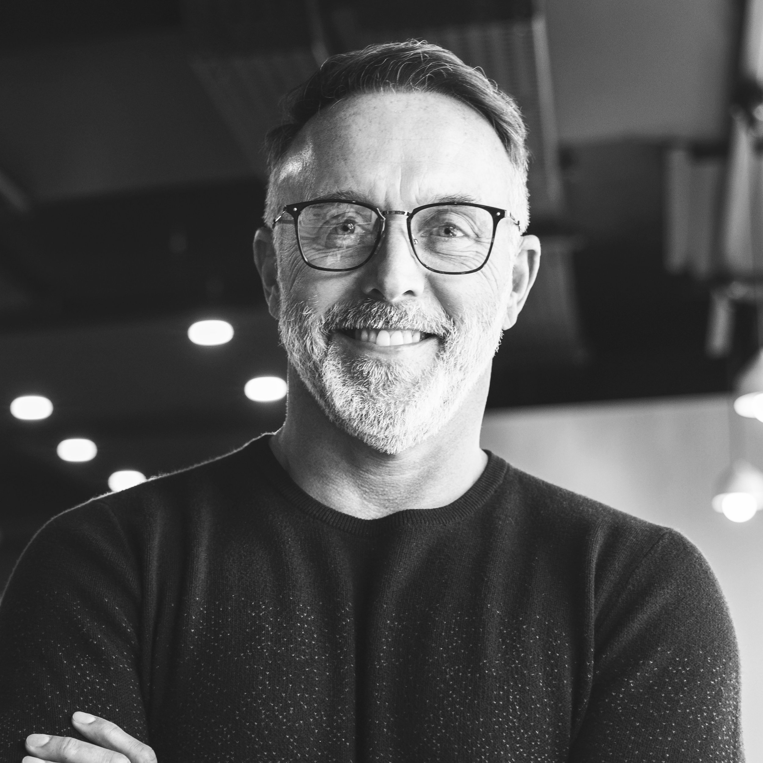 Black and white photo of a smiling middle-aged man with glasses, a beard, and short hair, wearing a black sweater, with a blurred indoor background featuring ceiling lights.