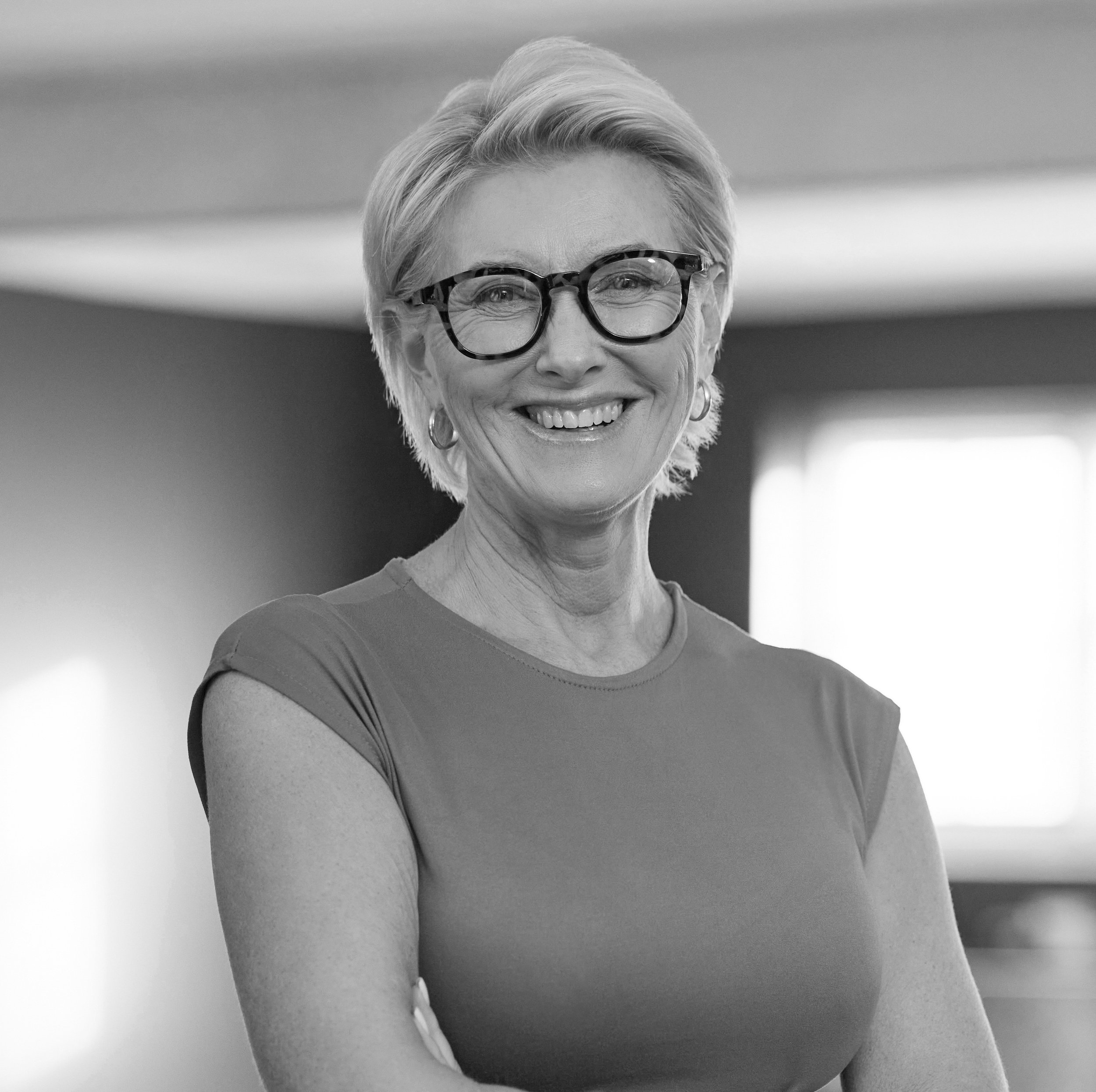A smiling woman with short, blonde hair, wearing glasses, earrings, and a short-sleeved top, standing indoors near a window.