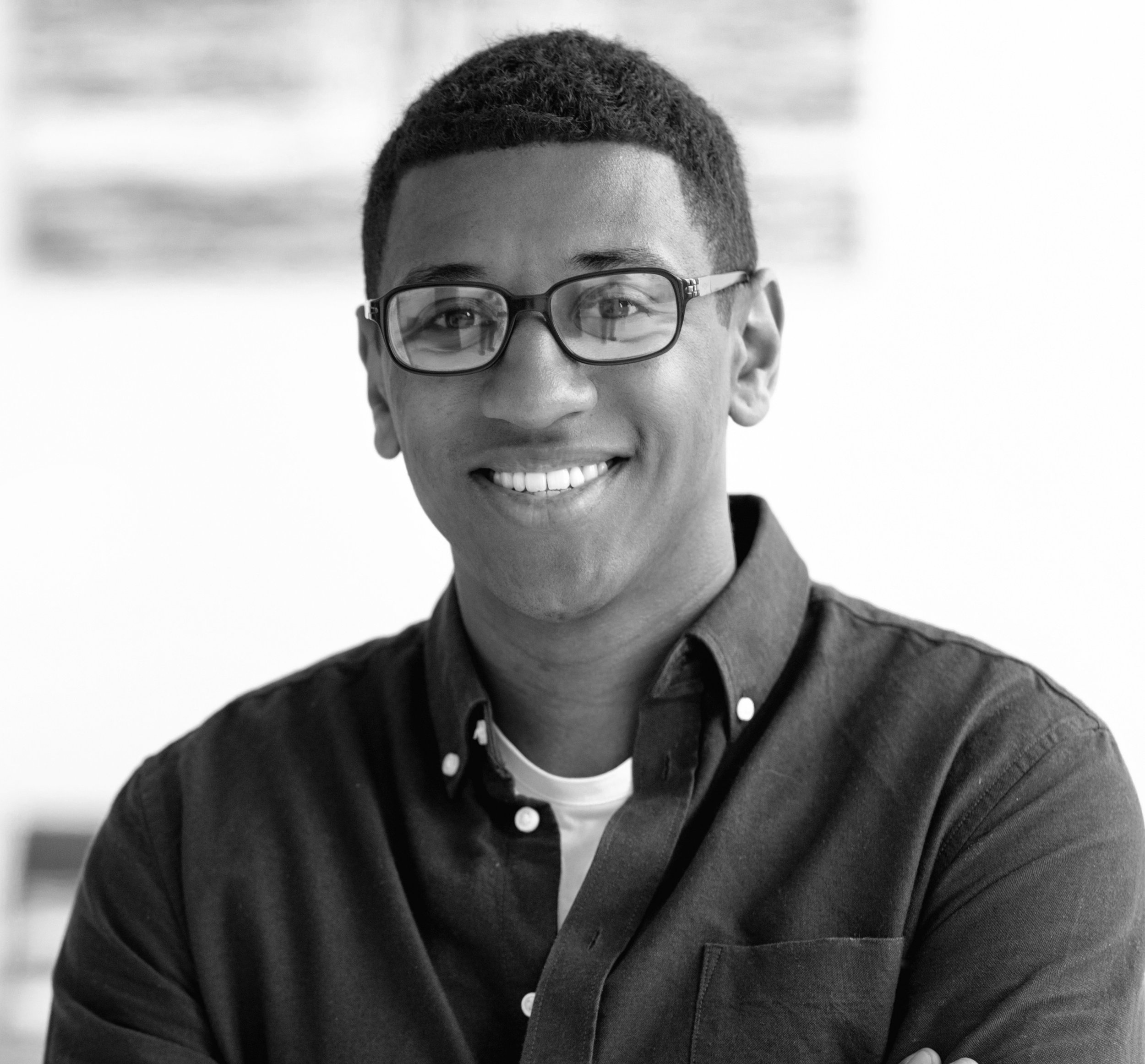 Black and white photo of a smiling young man with glasses, wearing a button-up shirt with a white T-shirt underneath, in an indoor setting.