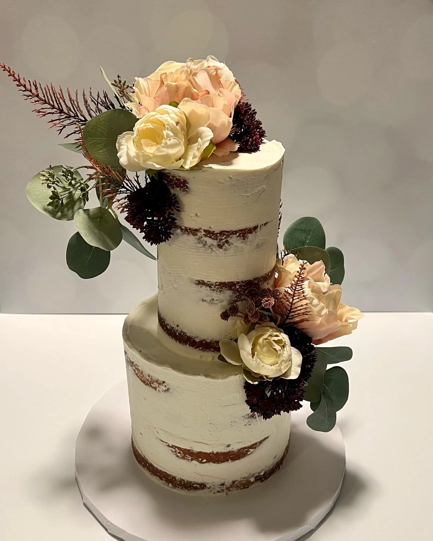 Two-tier semi-naked wedding cake with white frosting adorned with cream and blush roses, dark flowers, and green eucalyptus leaves, placed on a white cake stand at Cape Batter in Plymouth MA.