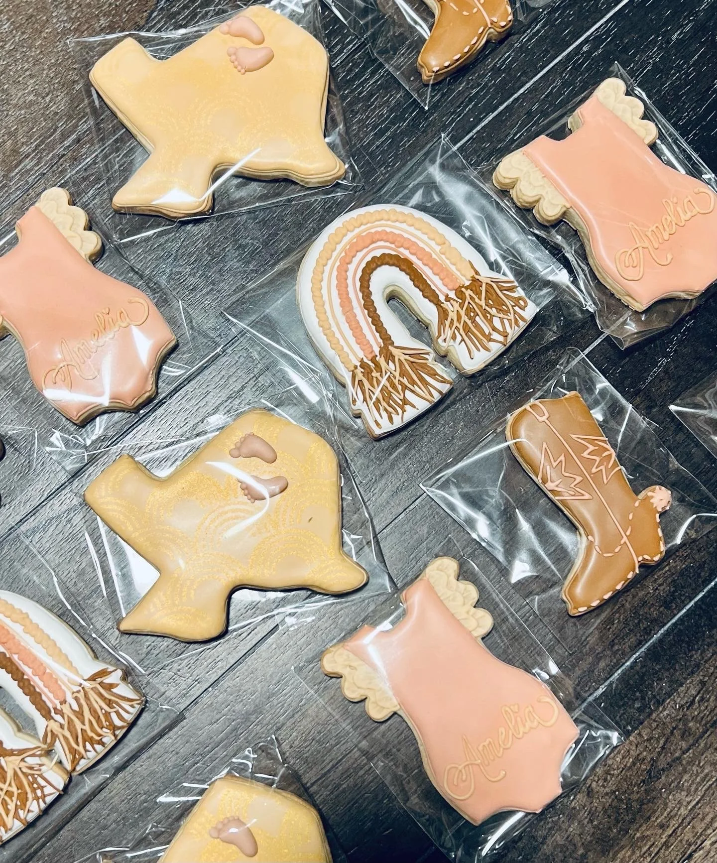 Decorated cookies shaped like cowboy boots, planes, onesies, and rainbows on a dark surface, each individually wrapped in plastic in Plymouth MA.