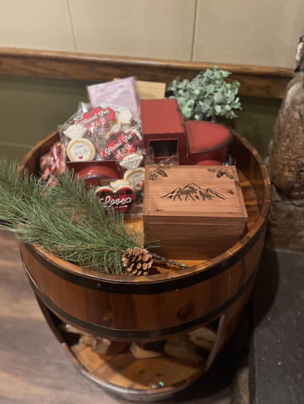 A wooden barrel end table displaying a variety of items including decorative cookies, a toy truck, a wooden box with mountain engravings, greenery sprigs, and a pine cone. The table is set against a rustic background with a stone texture on one side.