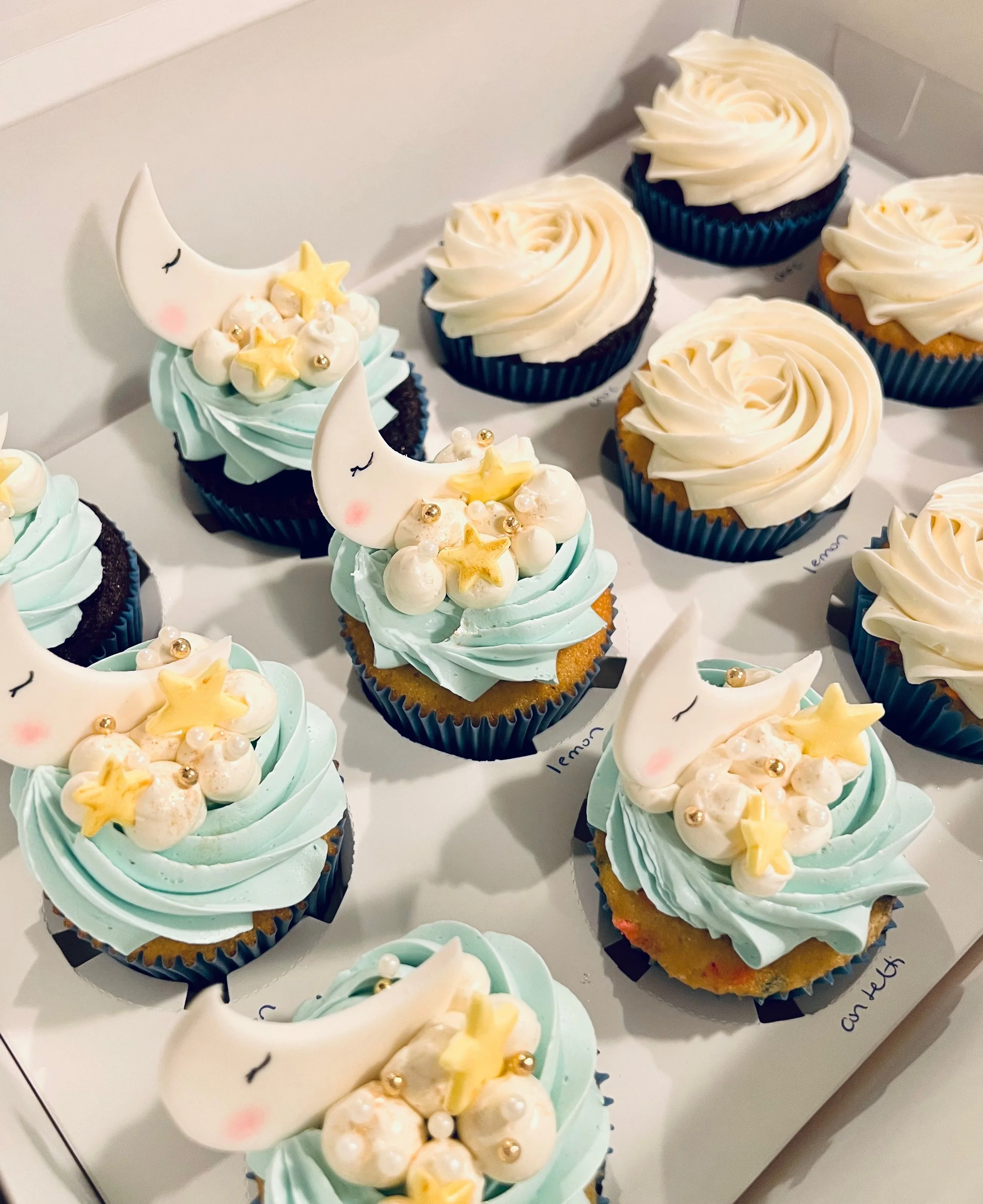Cupcakes with blue and white frosting, adorned with crescent moon and star shapes, placed in a box in Plymouth MA.