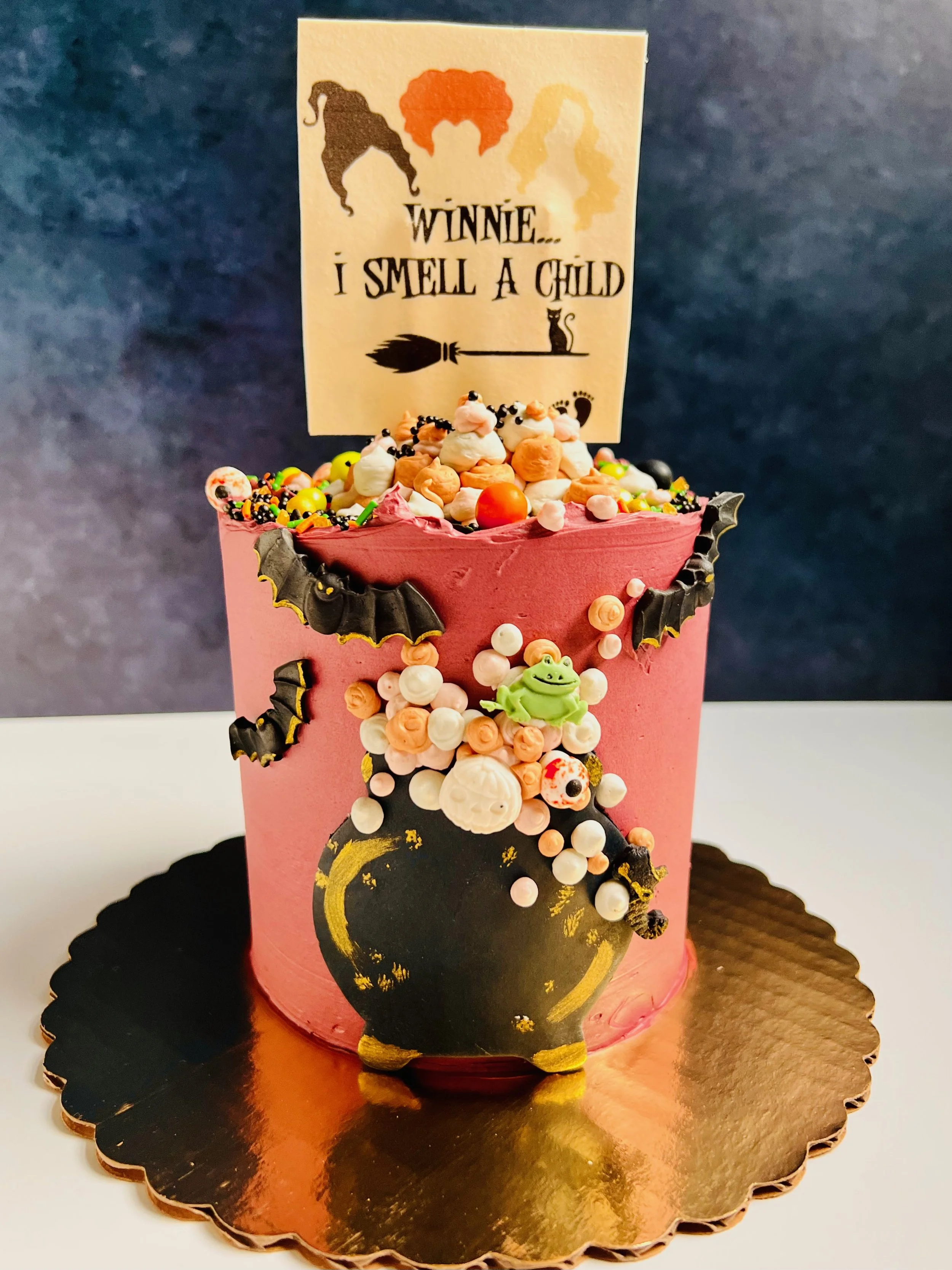 Halloween-themed cake with a pink frosting, colorful sprinkles, and a fondant cauldron design, topped with a sign featuring three silhouettes and the phrase 'Winnie... I smell a child.'