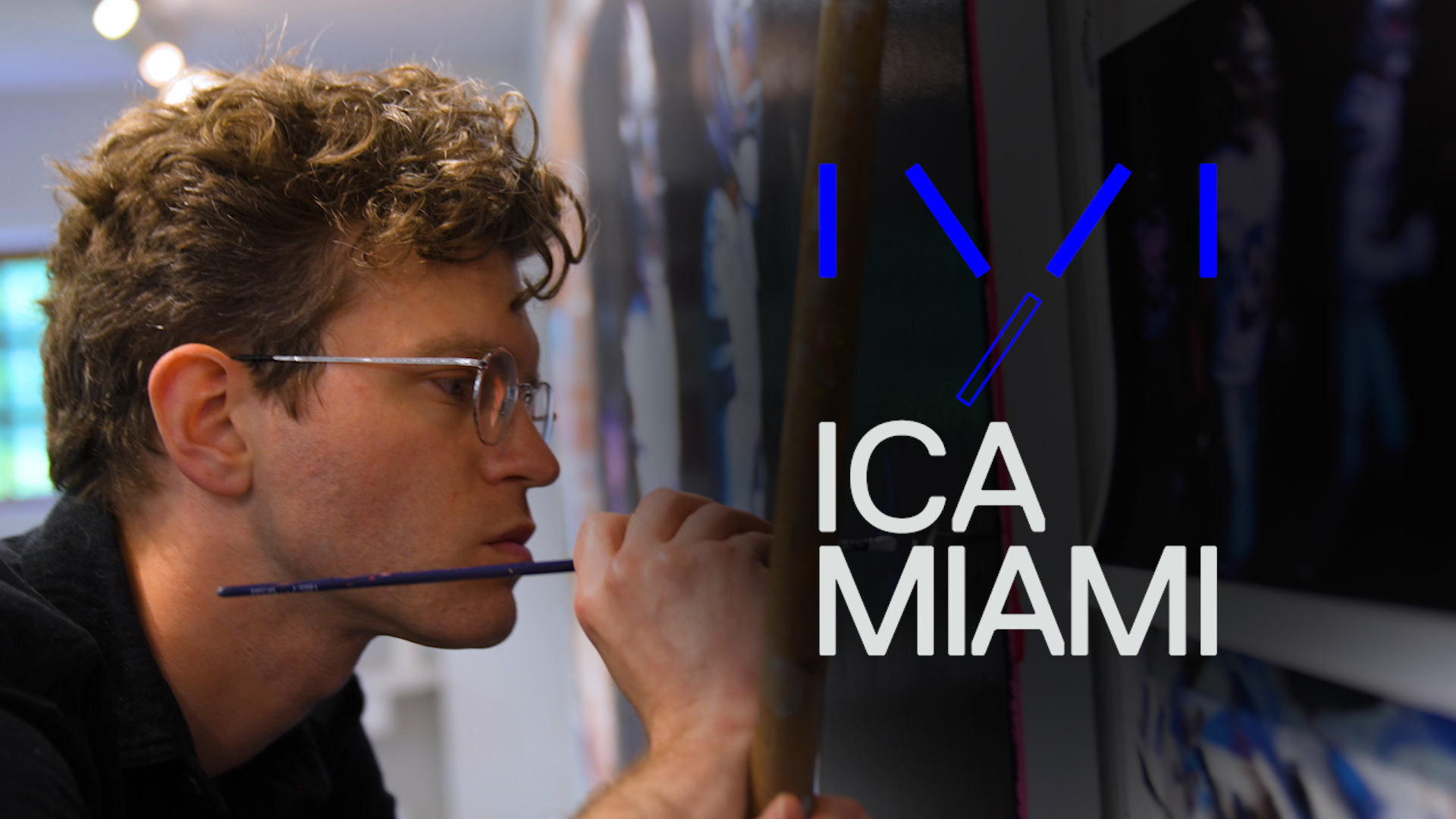 Close-up of a person painting with a brush near their mouth, wearing glasses, next to a wall with the text 'ICA Miami.'