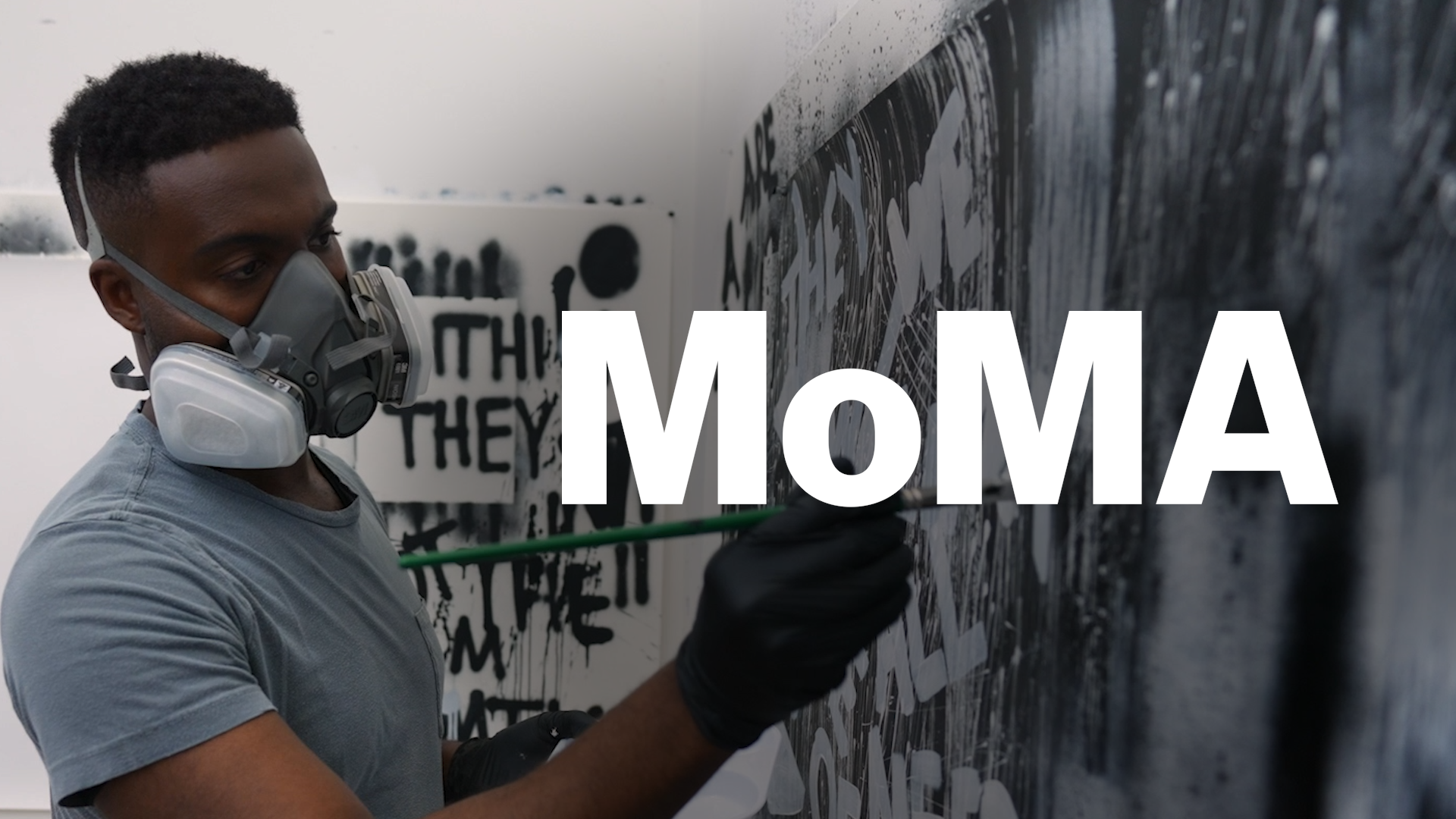 Artist wearing a respirator mask painting a black and white mural with text, featuring the word MoMA prominently displayed.