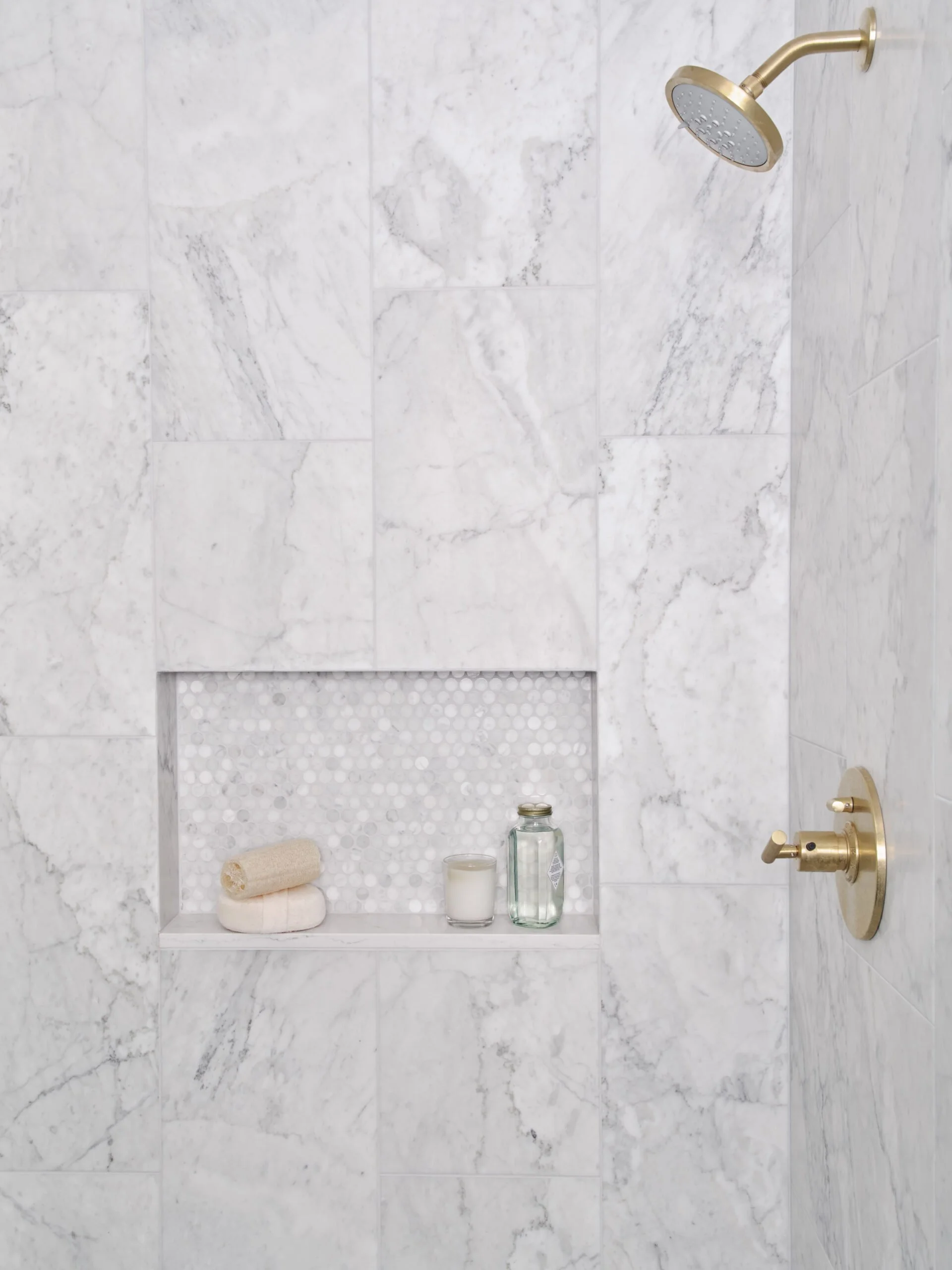 A marble shower wall with a built-in niche holding towels, a candle, and a glass bottle, with a gold showerhead and control valve.