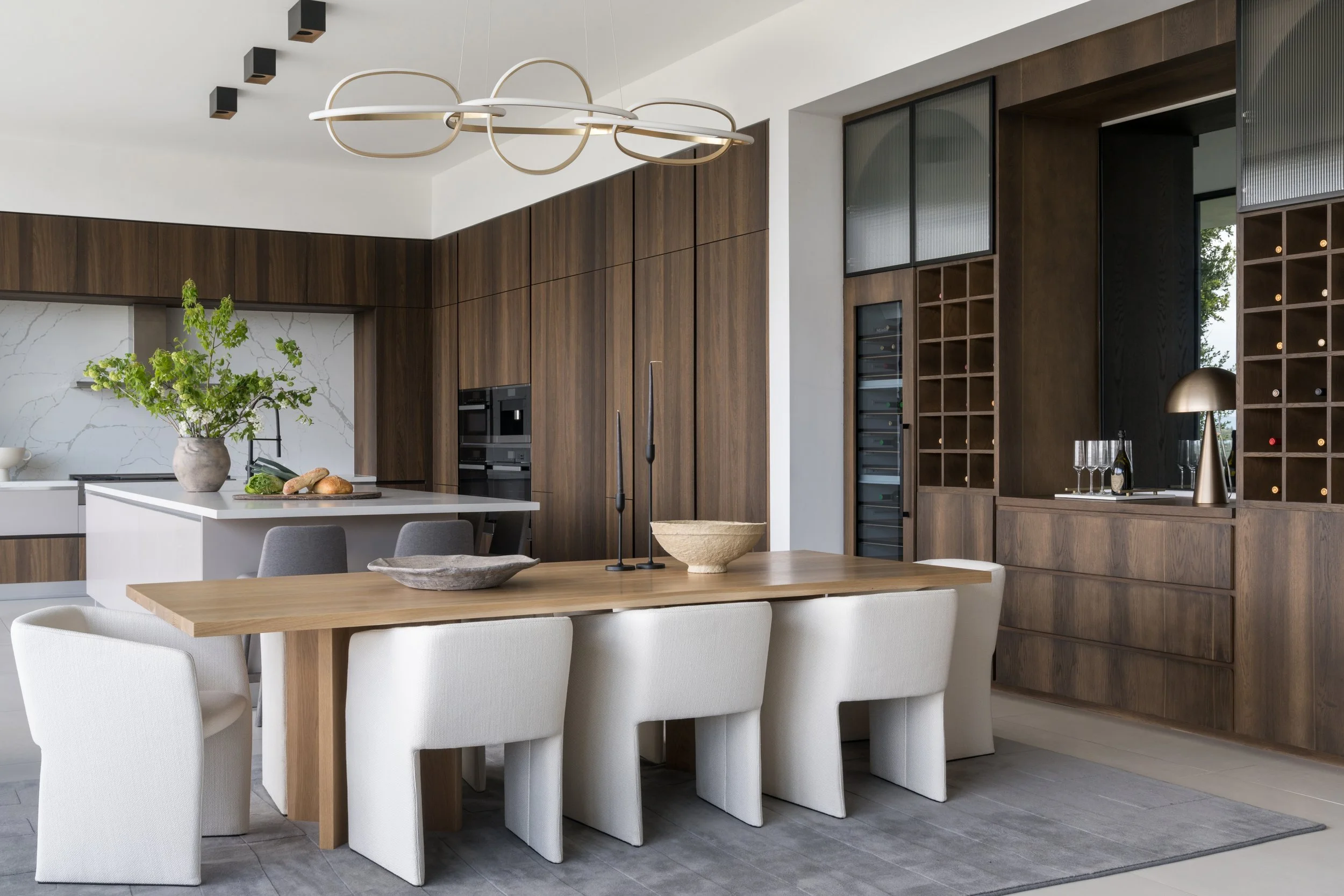 Modern kitchen with wooden cabinets, white marble backsplash, kitchen island with greenery, a wooden dining table with white and gray chairs, and a decorative light fixture overhead.