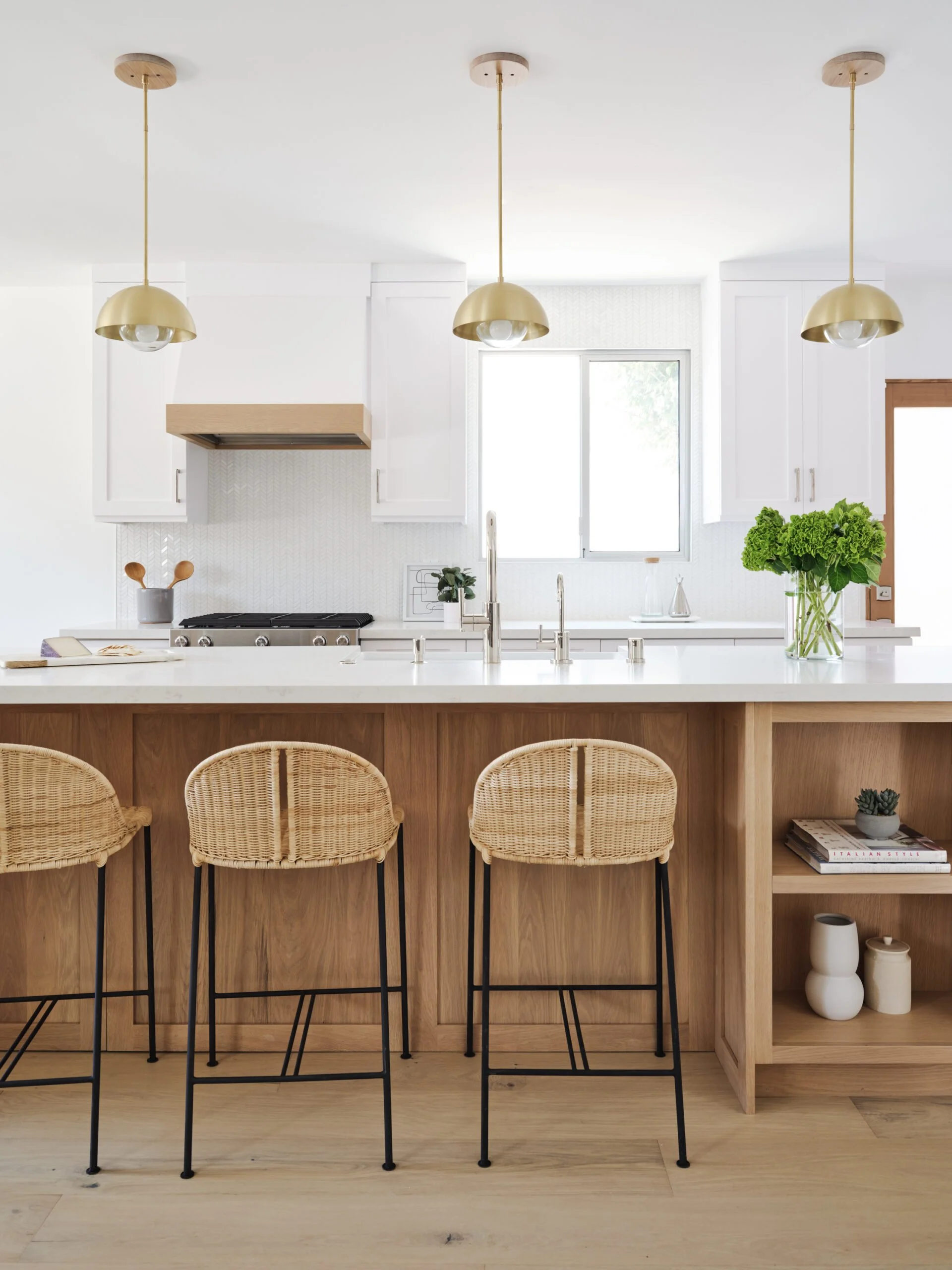 Modern kitchen with white cabinets, a wooden island with three wicker bar stools, pendant lights, and a window with a view outside.