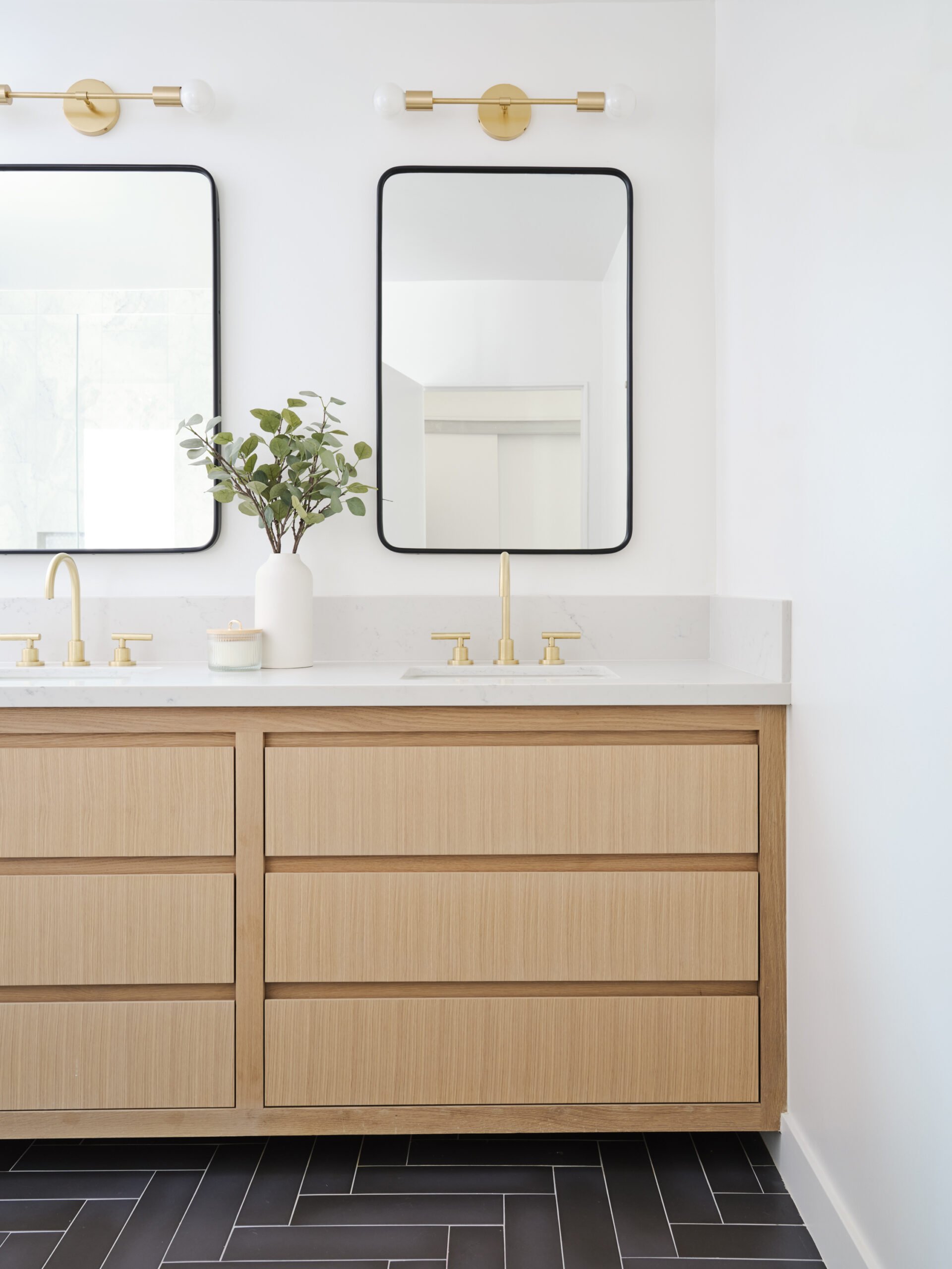 Bathroom vanity with wooden drawers, white countertop with two gold faucets, a white vase with green leaves, two rectangular mirrors with black frames, and a gold light fixture