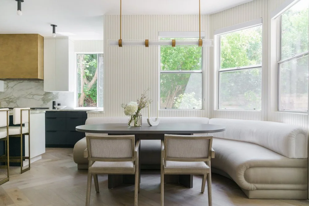 Bright dining area with a large white curved sofa, round black table, two beige chairs, a vase of white flowers, and large windows showing green trees outside.