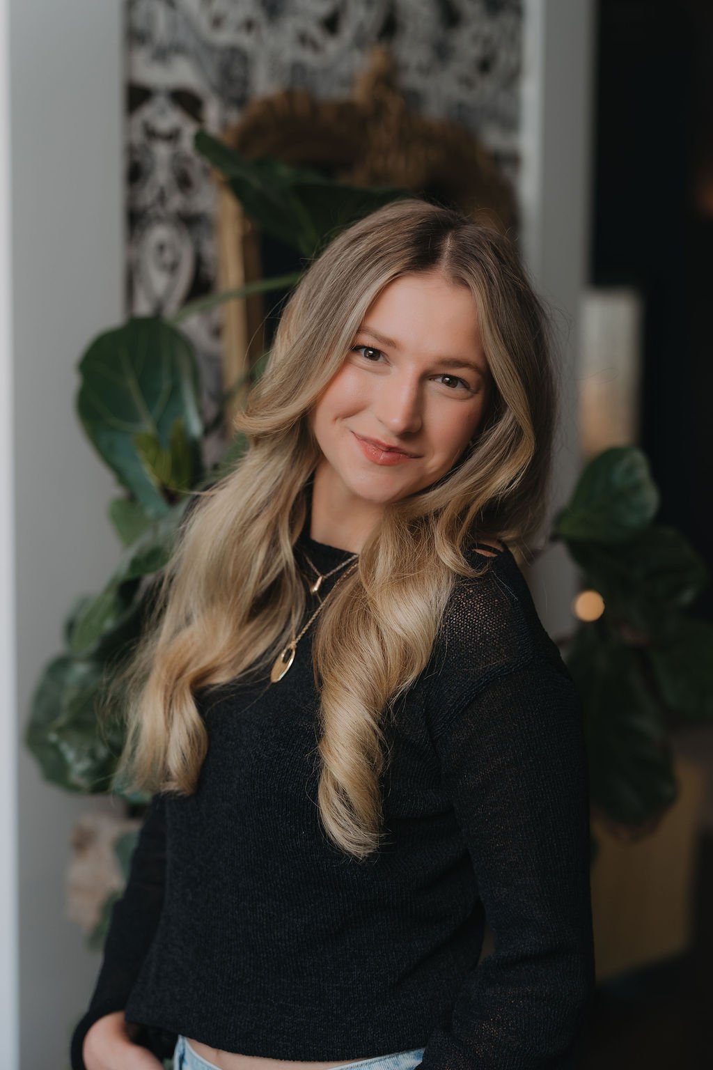 A young woman with long blonde hair, smiling, wearing a denim jacket and layered necklaces, in a cozy indoor setting with plants and warm lighting.