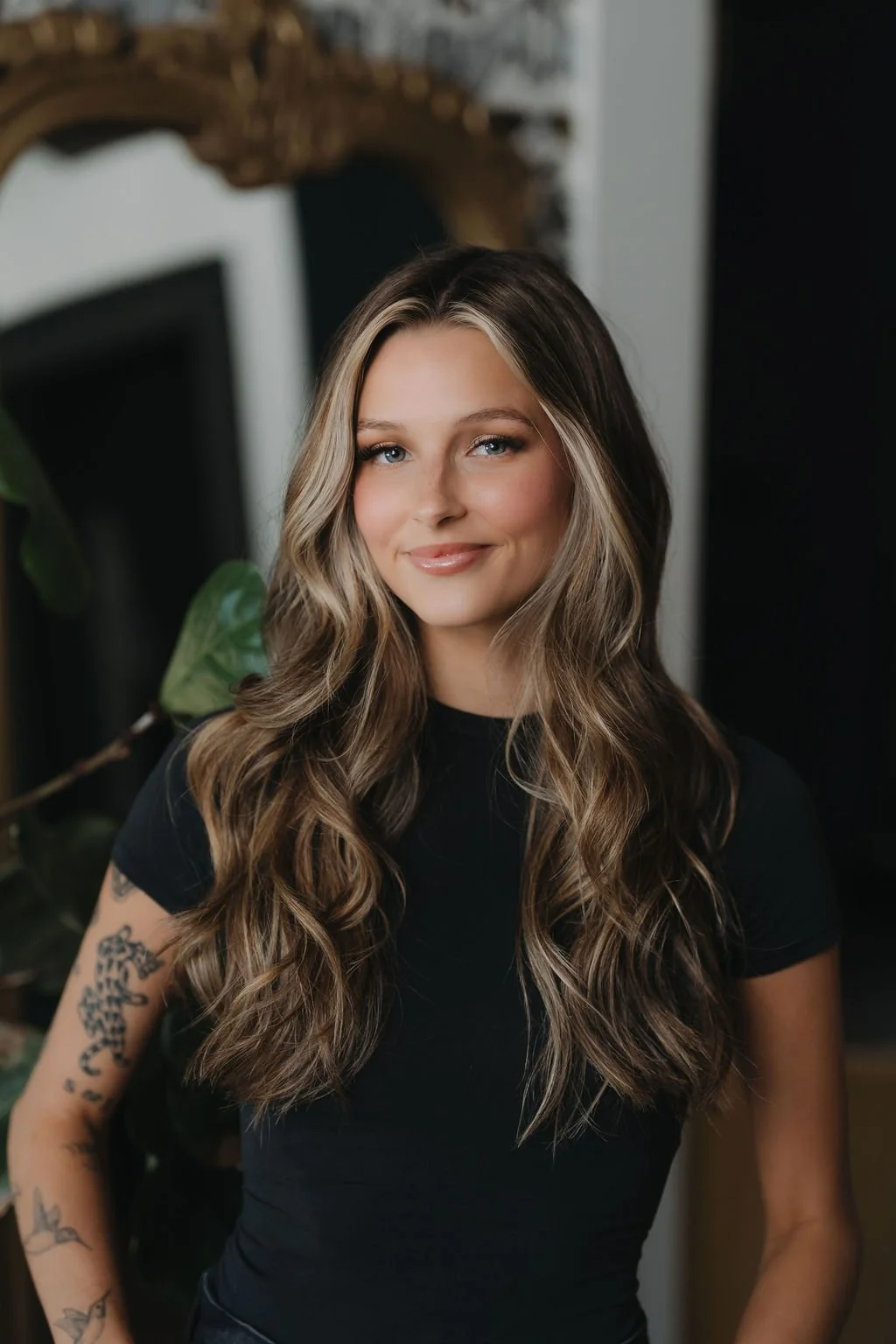 A young woman with long, wavy brown hair and blue eyes sitting in a cozy, dimly-lit indoor space, wearing a white off-shoulder sweater, smiling softly at the camera. Esthetican in Greenville.
