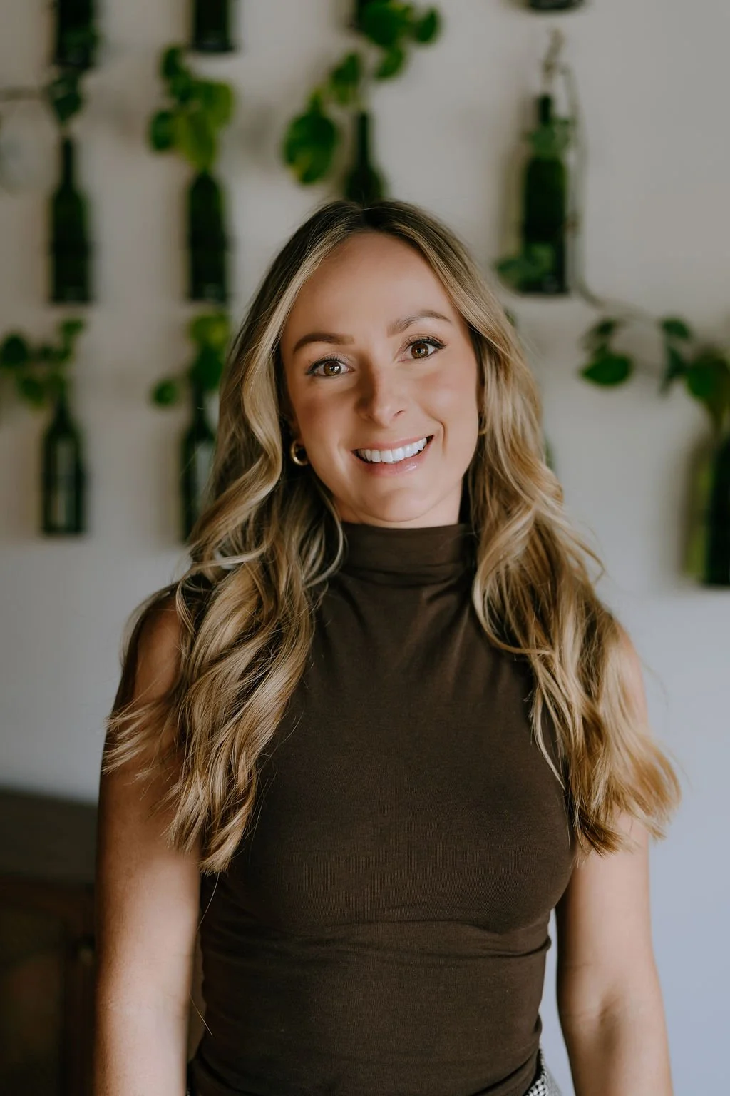 A woman with long brown hair, smiling, wearing a green patterned blouse and light-colored pants, standing indoors against a white wall and wooden floor. Spray Tan artist in Greenville.