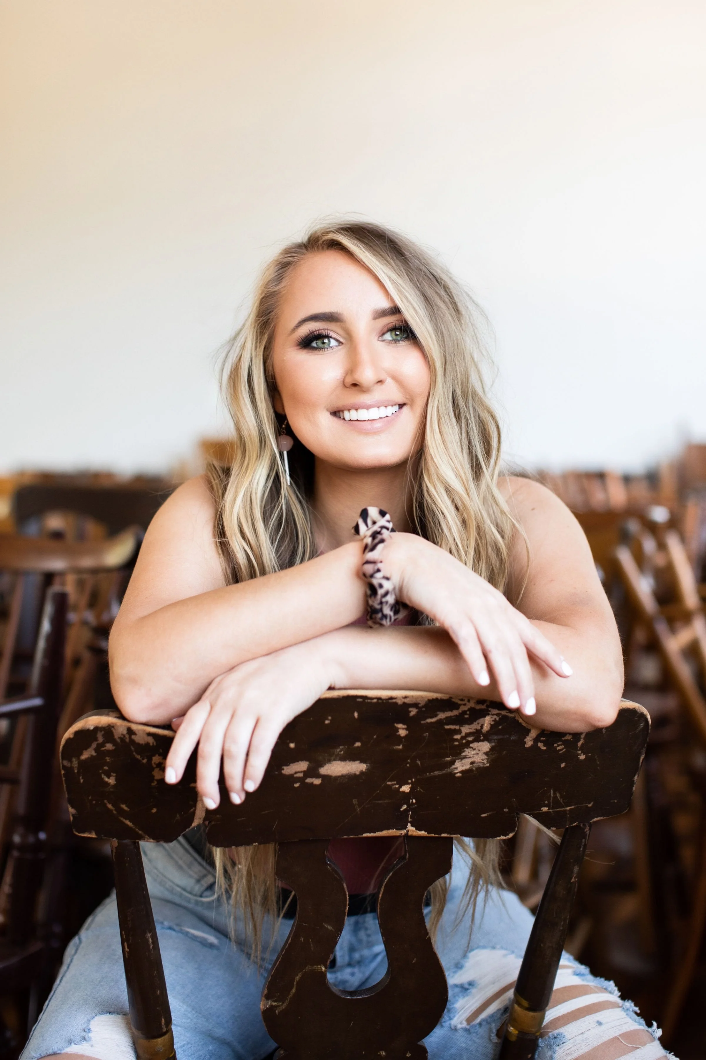 A young woman with long blonde hair, smiling, sitting in a wooden chair with her arms resting on the backrest, in a room with stacked chairs in the background.