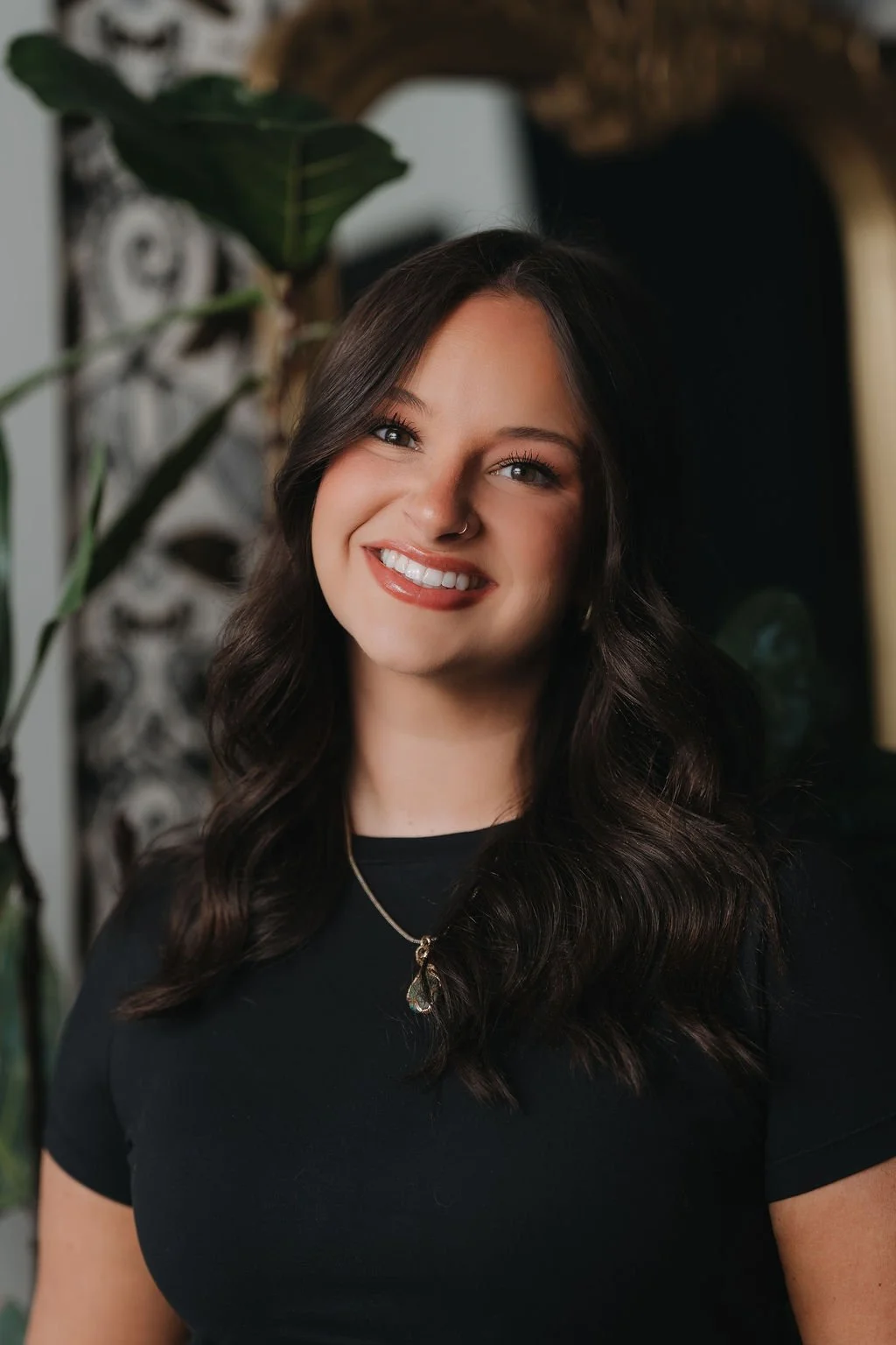 Young woman with dark wavy hair, smiling, wearing a beige sleeveless top, black skirt, gold earrings, and a gold necklace with a green pendant. Hairstylist in Greenville.