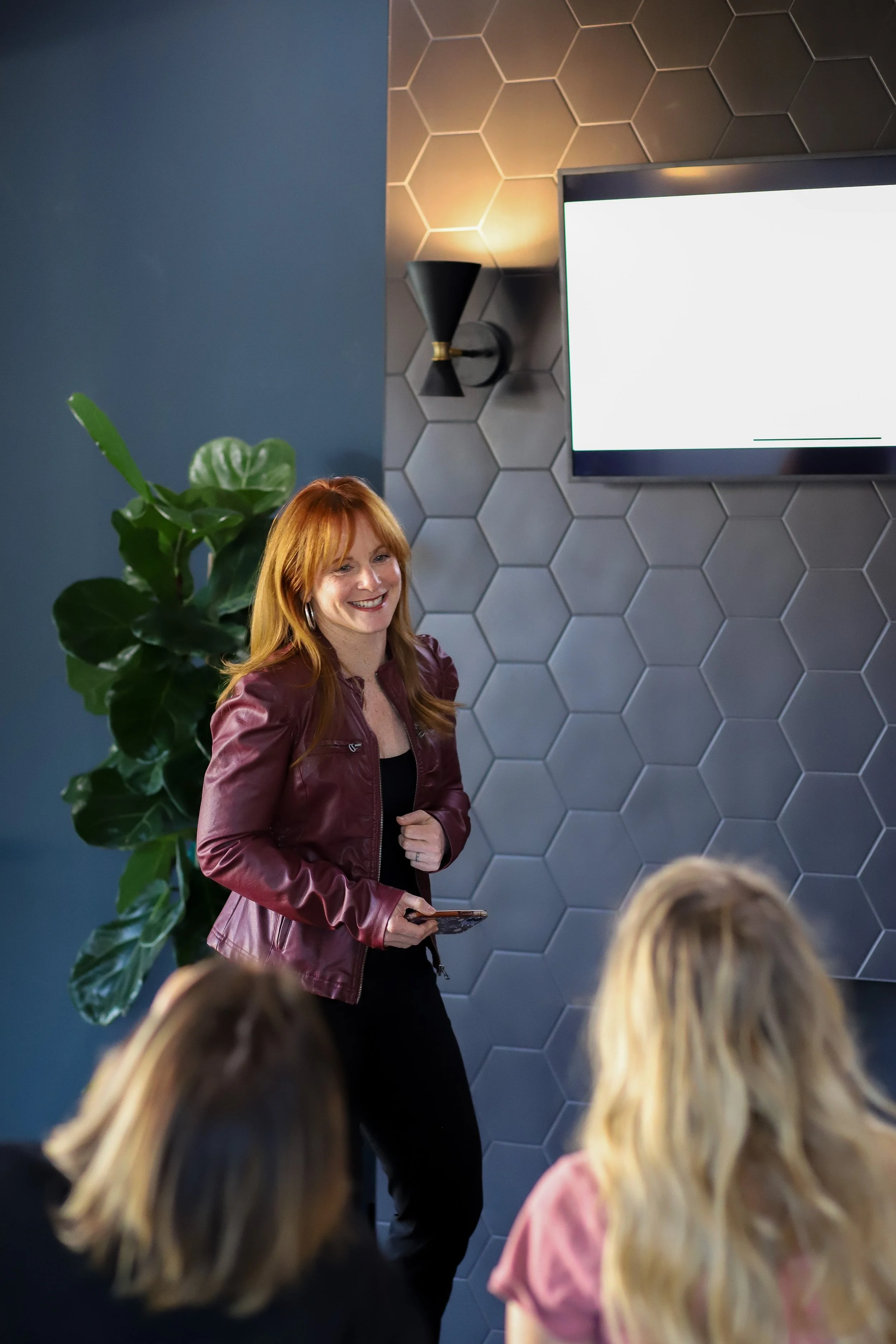 A woman with red hair, wearing a burgundy leather jacket, stands in front of a group, smiling during a presentation in a modern room with hexagonal wall tiles, a large TV screen, and a large green plant.