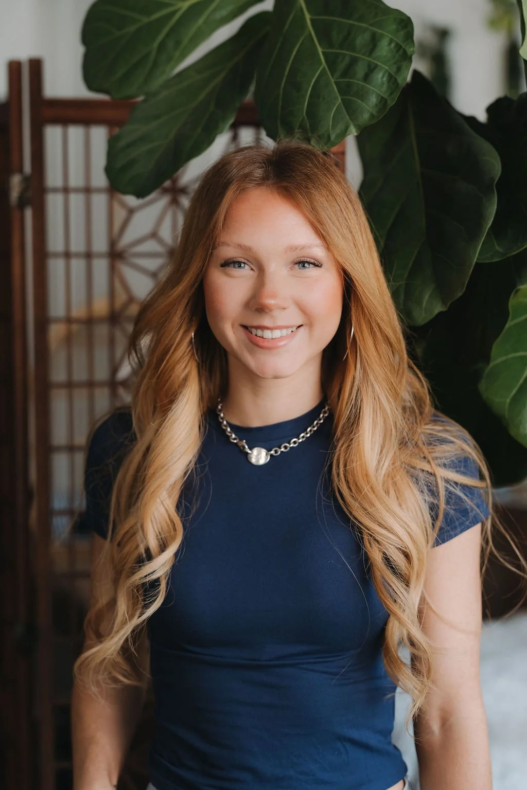 Young woman with long red hair smiling in a black shirt, with green plants in the background. Spray Tan artist in Greenville.