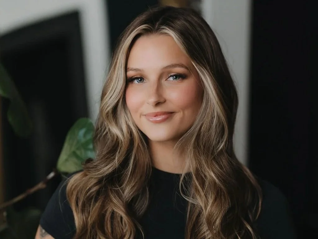 Portrait of a woman with long wavy hair, smiling, in a black top, with a blurred indoor background.