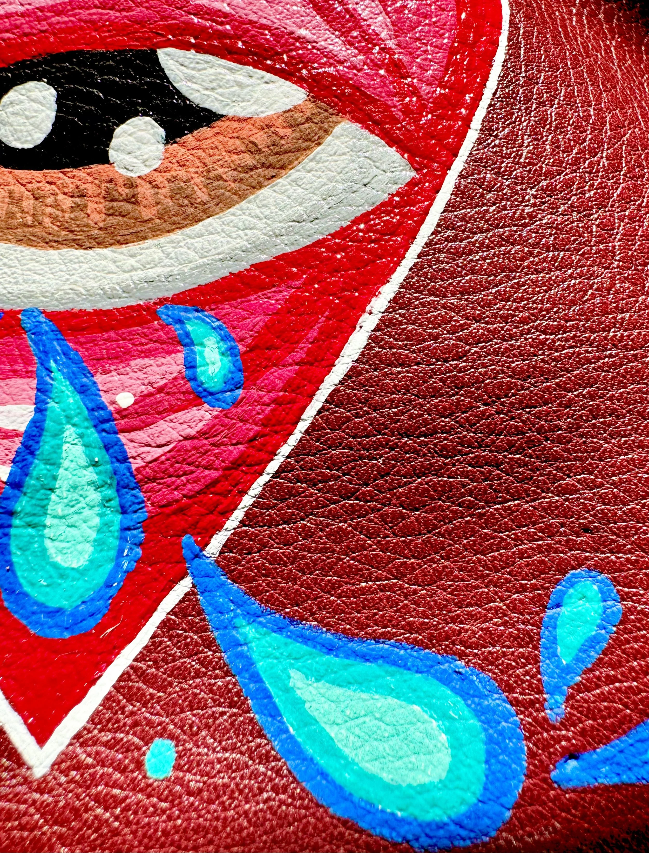Close-up of a colorful painted design on textured leather or similar material, featuring blue teardrops and a section of a face with eyes, nose, and red lips.