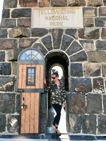 A woman standing at the entrance of Yellowstone National Park, under a sign, holding a door open in front of a stone wall.