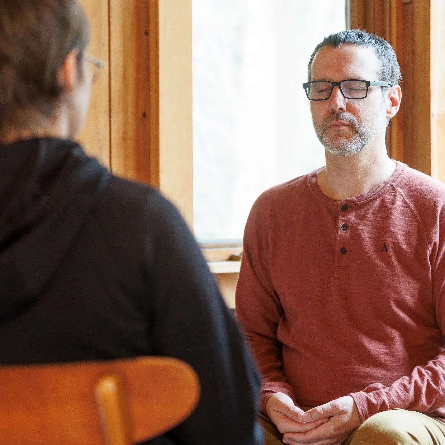 A man with glasses and a beard, meditating indoors with his eyes closed, sitting in a relaxed position near a window.