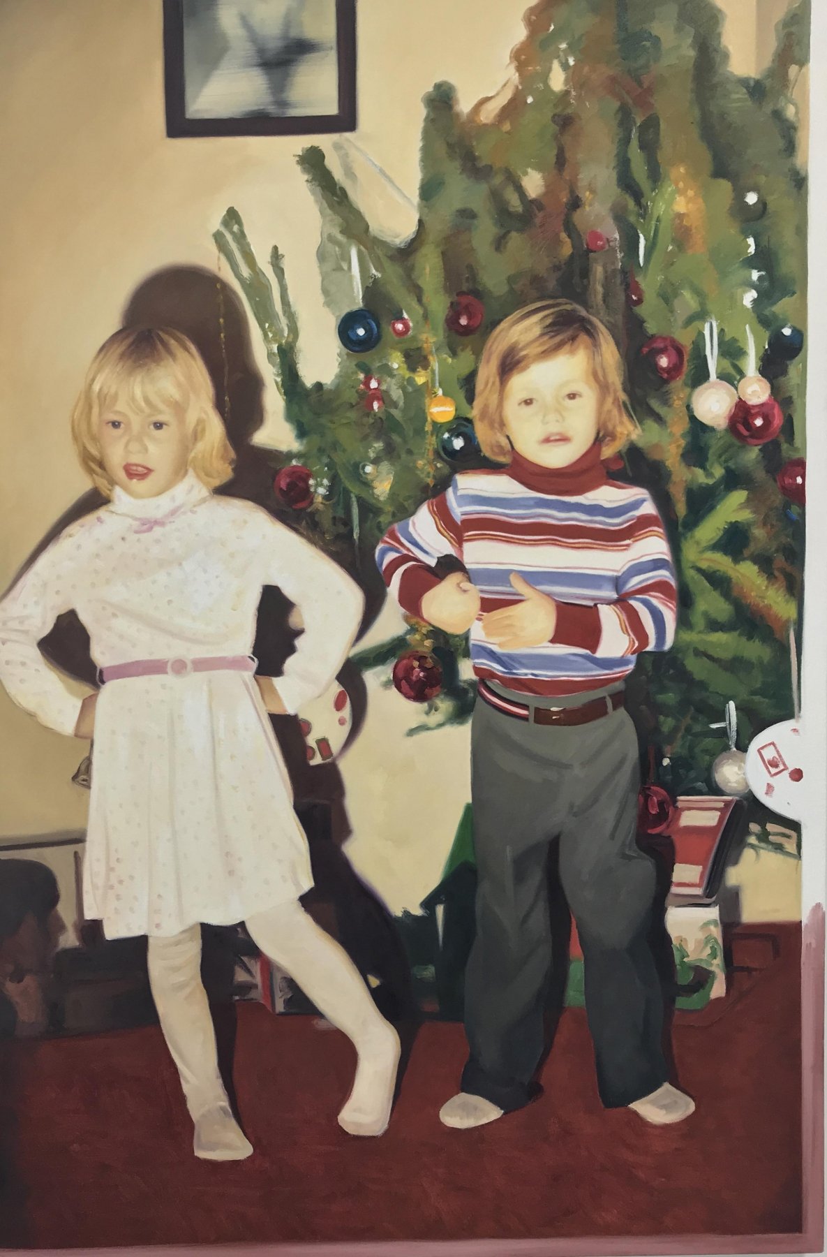 Two children posing in front of a decorated Christmas tree indoors, with one wearing a white dress and the other in a striped shirt and pants.
