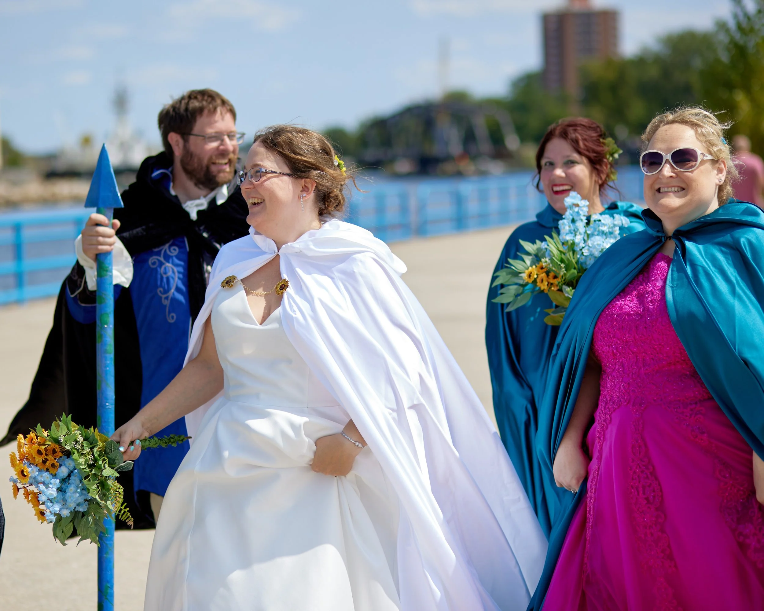 A group of four people dressed in fantasy costumes, standing outdoors by a waterfront, smiling and holding flowers, with a blue railing and buildings in the background.
