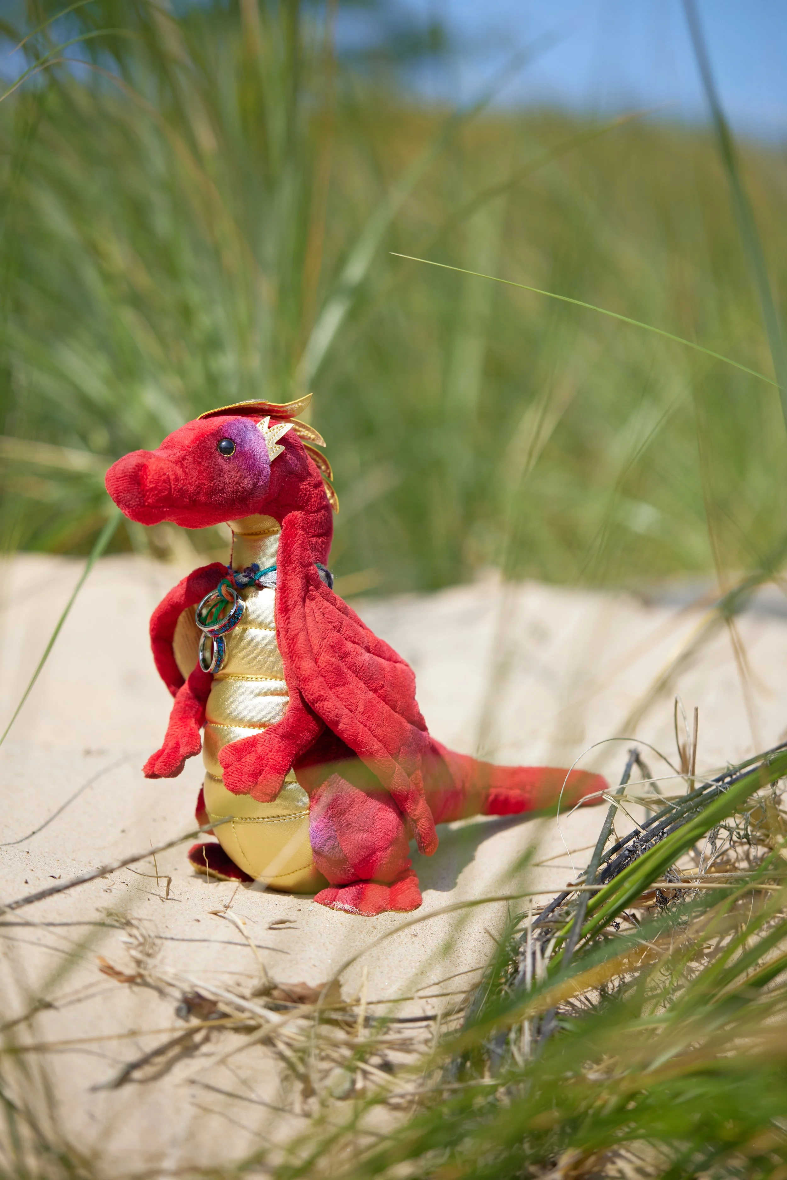 A plush toy dinosaur with a red body, cream-colored belly, and gold accents, wearing jewelry, sitting on sandy ground among green grass and plants.