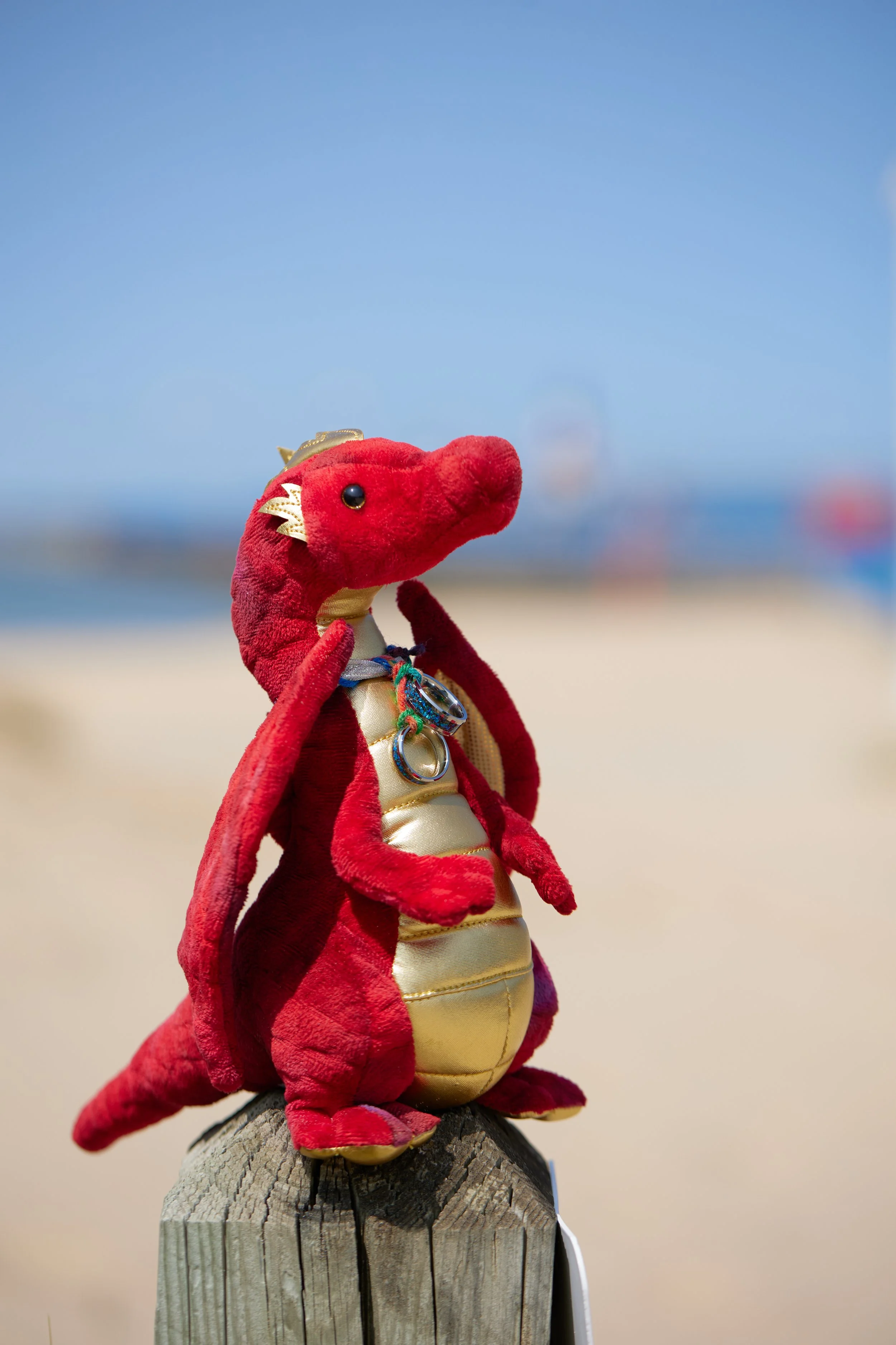 Red plush toy dinosaur with gold accents, wearing jewelry, on a wooden post at the beach.