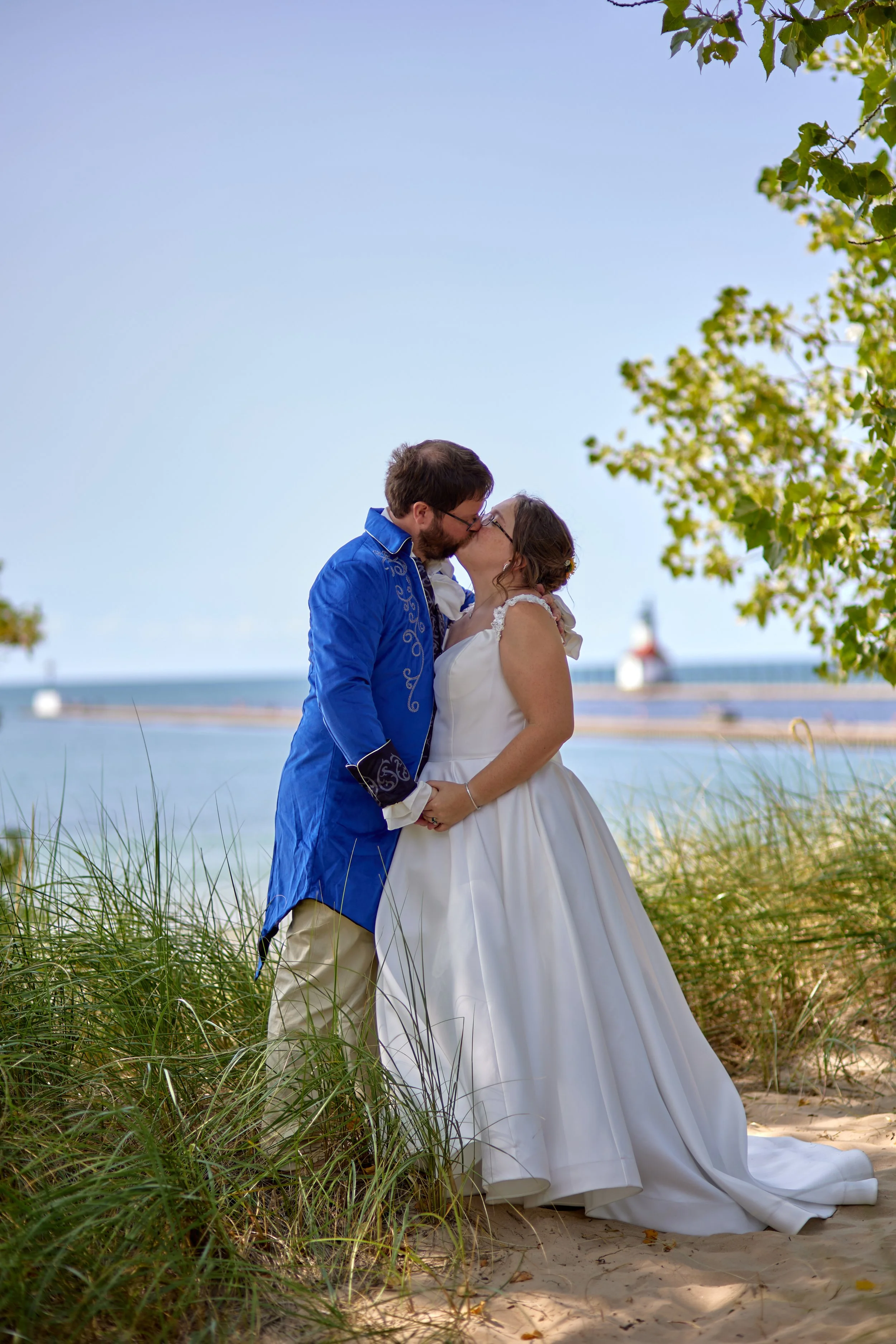 A newlywed couple sharing a kiss on a beach, with the ocean and a lighthouse in the background, surrounded by green foliage.