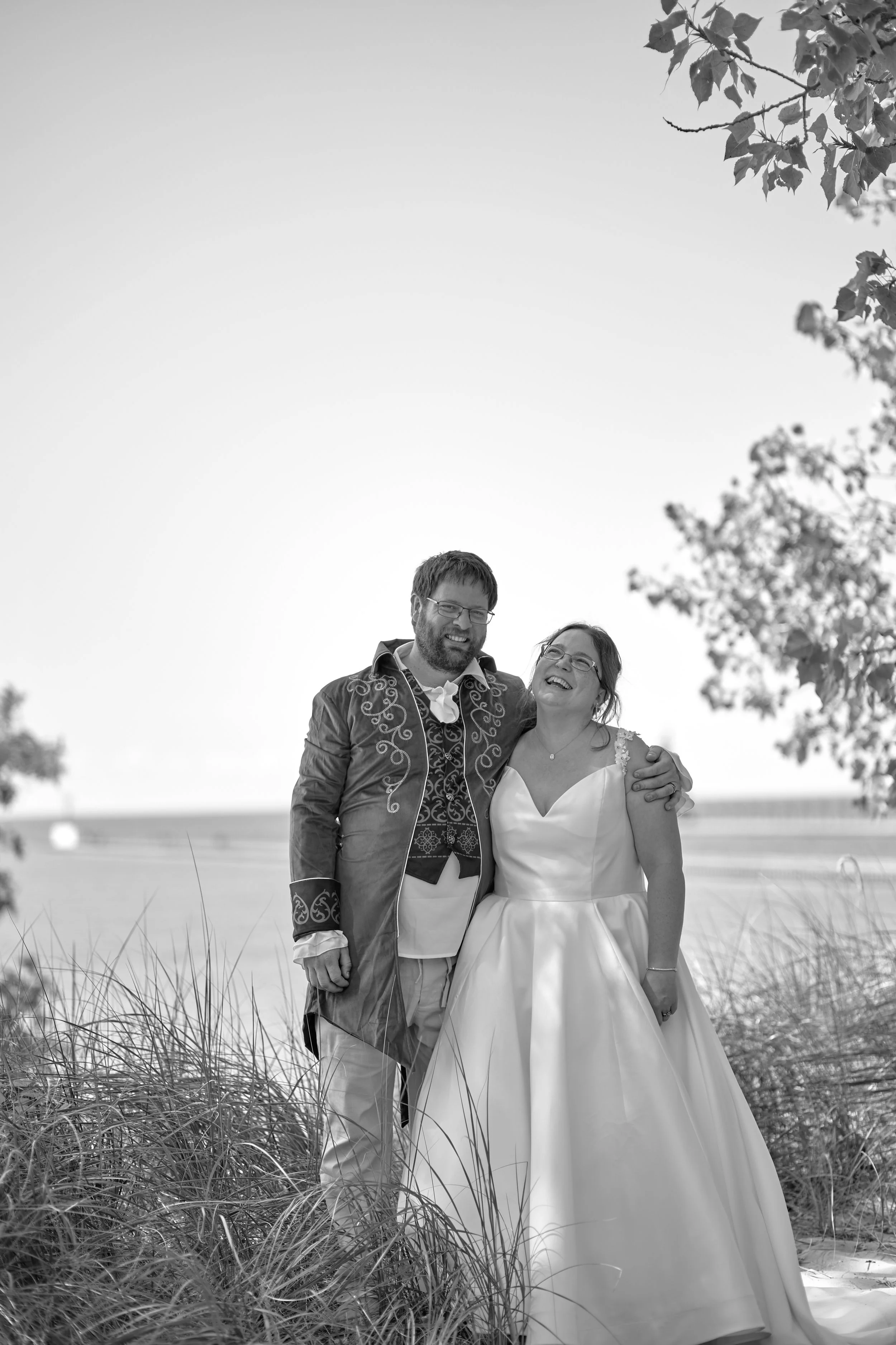 A smiling couple in wedding attire standing outdoors among sand and grass, with a body of water and trees in the background, captured in black and white.