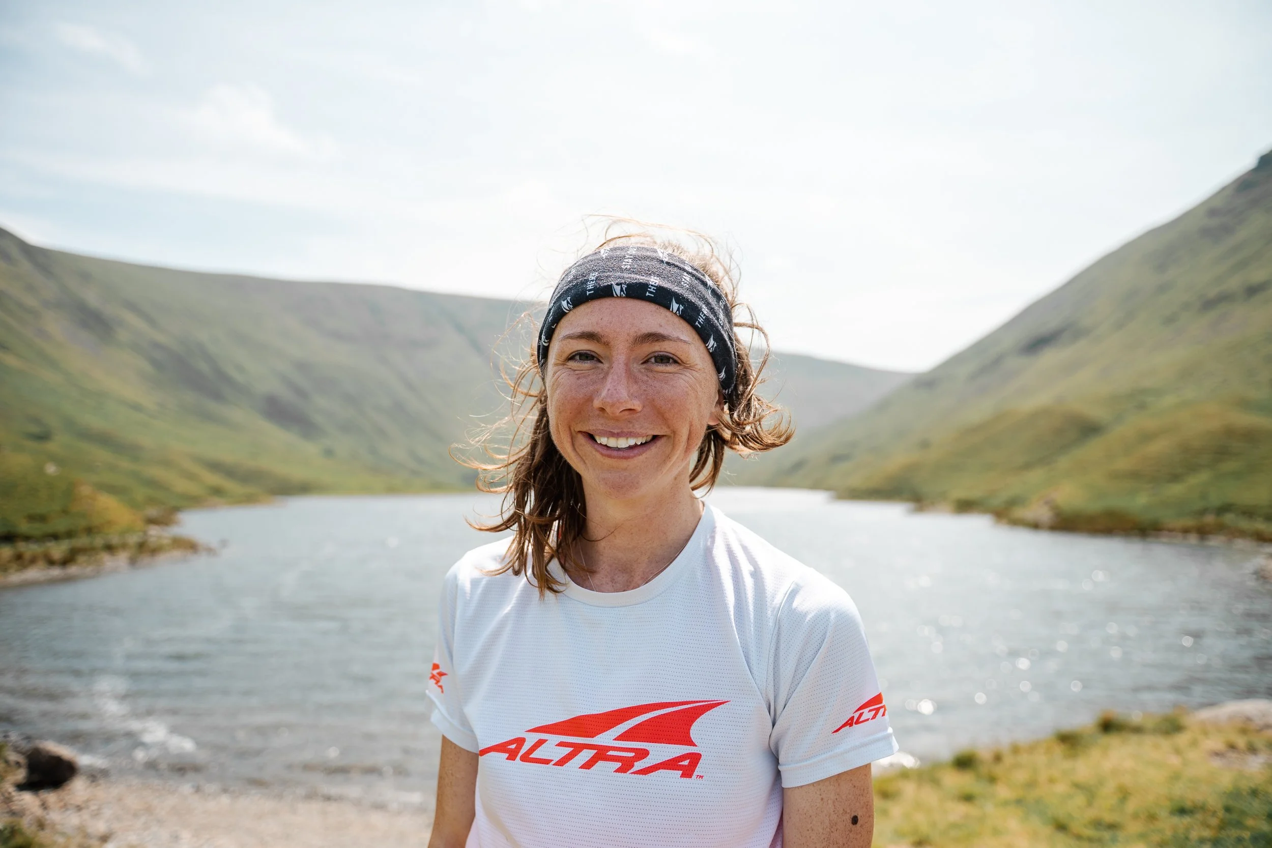 Woman wearing a white t-shirt and a headband smiling in front of a sunny lake