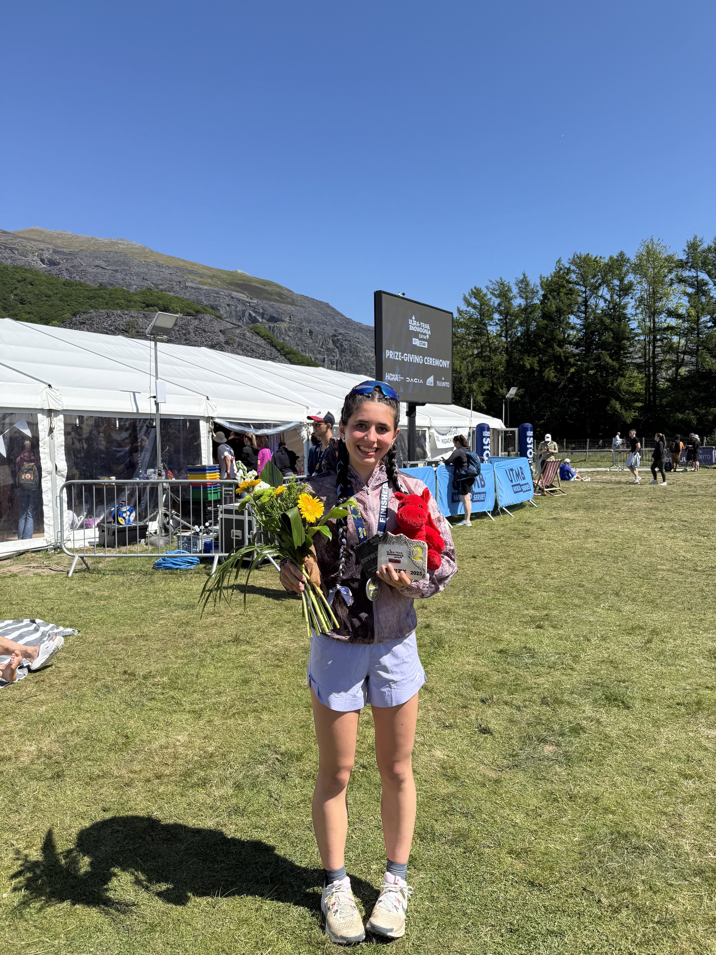 A young woman wearing sports clothing is holding a trophy and a bouquet of flowers on a sports field